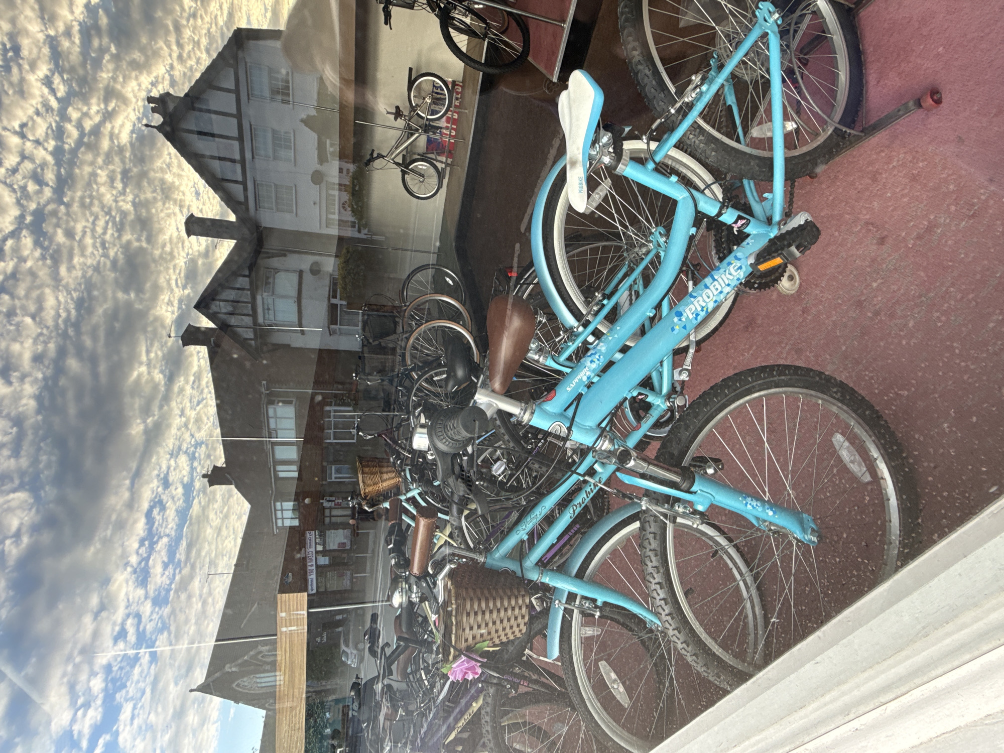 The image shows a shop window in Great Yarmouth, United Kingdom, displaying several bicycles. The bicycles are various models and colors, with one prominently being a light blue "Probike" model. The scene is layered with reflections. In the reflection, buildings are visible across the street, with a sky full of scattered clouds. The background inside the shop appears to be a red or maroon color, likely part of the shop's interior.