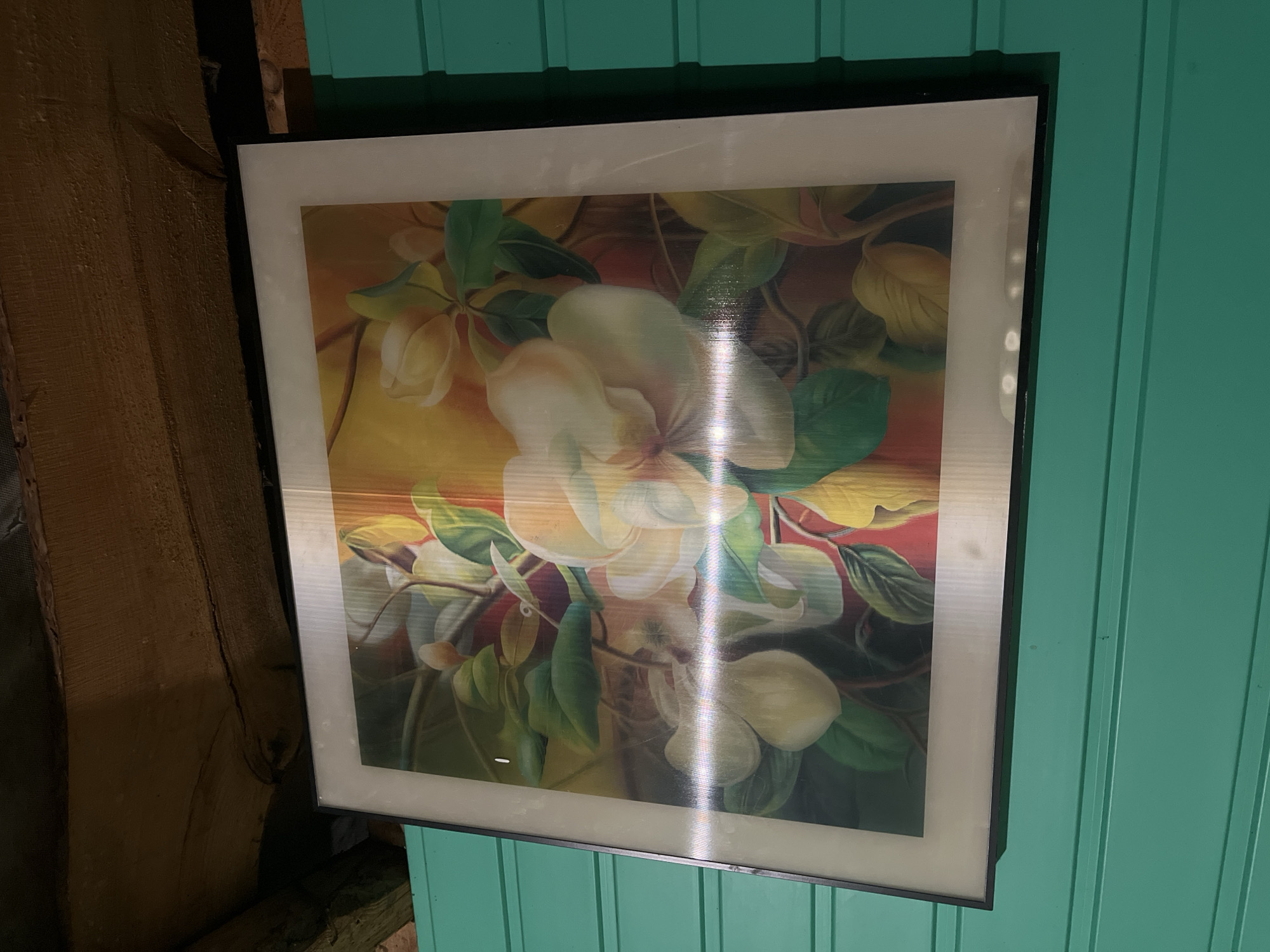 A framed floral painting hangs on a teal-colored, horizontally paneled wall. The painting features large white and light yellow blossoms with green leaves against a background that transitions from yellow at the top to reddish-brown at the bottom. The artwork is framed with a thin black border and a wider white mat. Above the painting, rough-hewn wooden beams form the ceiling, suggesting an interior space. The overall scene is static and depicts a decorative element within a rustic setting. This is a photograph taken in Abzakovo, Russia.