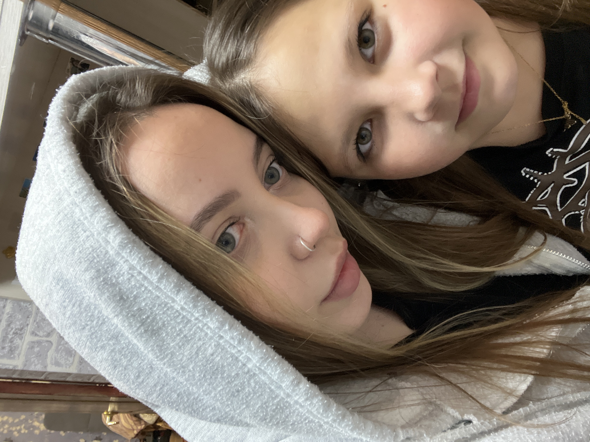 Two individuals, presumably female, are close together, facing the camera. One is wearing a white hooded sweatshirt. The younger one has dark hair and is wearing a necklace with a black shirt. The scene appears to be indoors. The location is Novopokrovskaya, Russia.