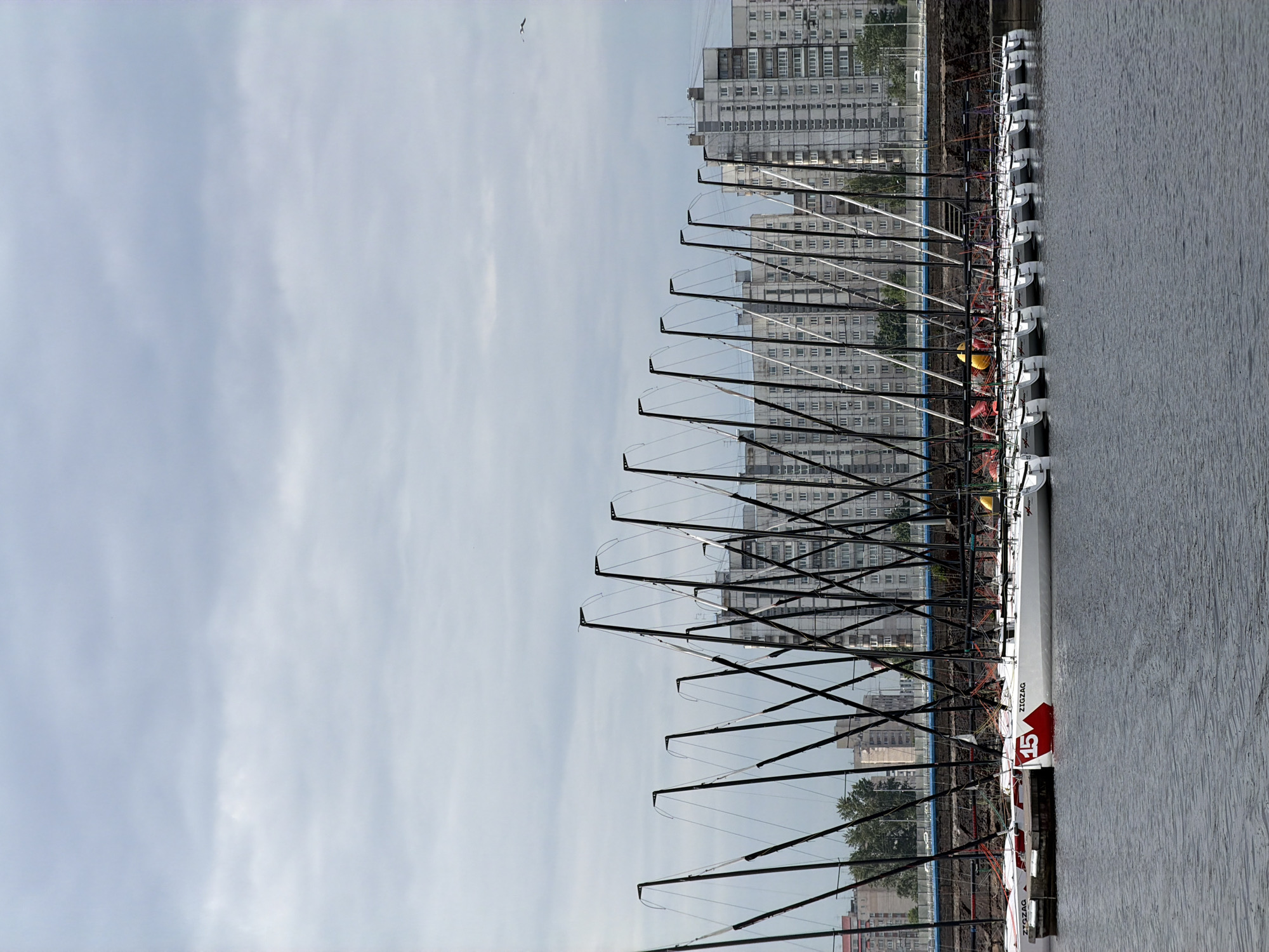 The image depicts a fleet of identical white sailboats docked in a harbor. Each sailboat features black masts with rigging and is lined up neatly along a pier. The boats are branded with the word "ZIGZAG" and one prominently displays the number "15" in a red shield. In the background, several large, grey apartment buildings rise against a cloudy sky. The water in the foreground is a calm, dark grey. The scene appears to be located in Saint Petersburg, Russia, given the architectural style of the buildings and the maritime setting.