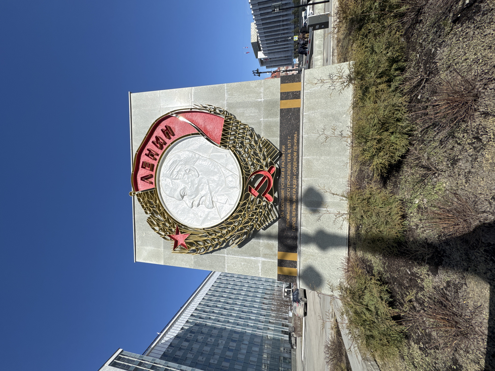 The image presents a forensic view of the Order of Lenin monument in Perm, Russia, under a clear, bright blue sky. The central subject is a large, grey, monolithic monument. Its primary feature is a prominent circular emblem with a white relief bust of Vladimir Lenin, facing left. This bust is encircled by a red banner displaying the Cyrillic word "ЛЕНИН" (LENIN) in gold lettering. Stylized golden wreaths frame the inner circle, topped by a red star and featuring a red hammer and sickle symbol at the base. Below this emblem, a dark plaque with white Cyrillic inscription confirms: "FOR GREAT SUCCESSES IN THE DEVELOPMENT OF INDUSTRIAL PRODUCTION IN 1971. THE CITY OF PERM WAS AWARDED THE 'ORDER OF LENIN'". The plaque is bordered by a horizontal black strip with two yellow bands.
The monument is set within a landscaped median in the foreground, characterized by dark soil and a mix of green coniferous shrubs and bare-branched deciduous plants. Bright sunlight casts distinct shadows, including that of the photographer, on the ground. To the immediate left, a large, multi-story modern building with a facade primarily composed of glass and light-colored panels reflects the sky. In the background to the right, another substantial building with a more traditional Soviet-era architectural style, featuring vertical elements and a lighter color, is visible; flags are perched on its roof. Several vehicles, including a light-colored SUV, are parked on paved areas behind the monument. A small group of individuals is walking on a paved pathway in the mid-ground to the right, near the second building, with a white and red automated barrier arm visible adjacent to them. The scene depicts a public square or street environment.