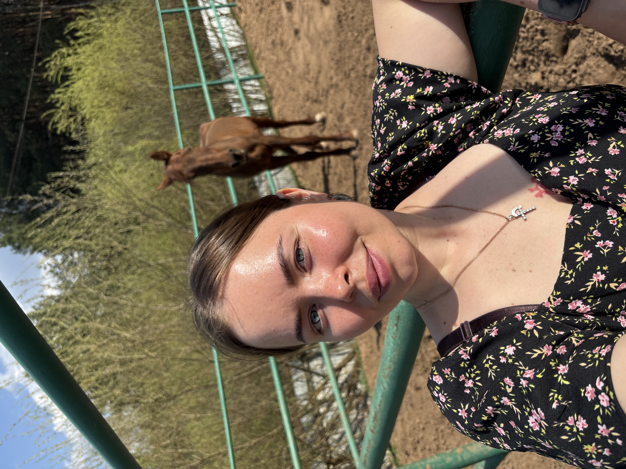 A woman is posing for a selfie in front of a green metal fence. Behind the fence, a brown horse is visible in a dirt enclosure. The woman is wearing a black floral dress and a silver necklace. The location appears to be rural, possibly a farm or ranch, and is not explicitly identified as Русский Вожой, Russia. The woman is holding the fence.