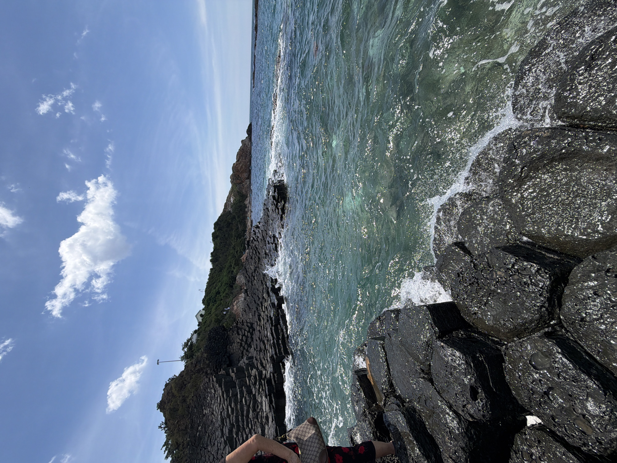 An eye-level, wide shot captures the coast of Phu Luong, Vietnam, on a bright, sunny day. The foreground is dominated by dark, hexagonal basalt rocks, some of which are wet from the incoming tide. A portion of a person's legs and a handbag are visible on the left, standing on the rocks. The sea is a clear, turquoise color with gentle waves breaking against the shore. In the middle ground, more of the distinctive basalt formations extend into the water. The background features a green, vegetated hillside with a few small buildings and a lamppost. The sky is a vibrant blue with scattered white clouds.