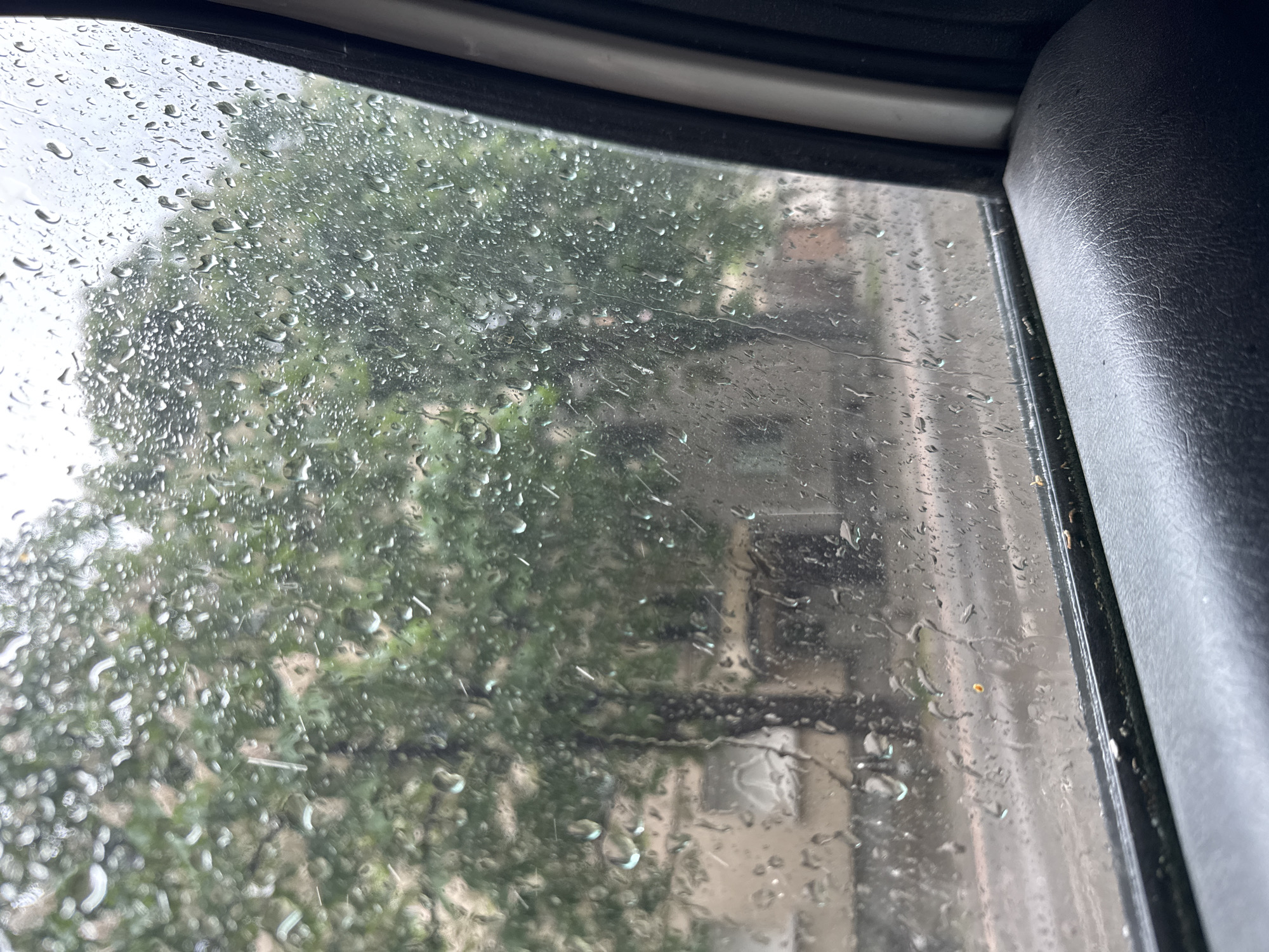 The image shows a view through a rain-streaked car window. Outside, trees with green leaves are visible, and a building is partially visible in the background. The building has windows and a facade. The car's interior, including the window frame and part of a seat, is visible on the edges of the frame.