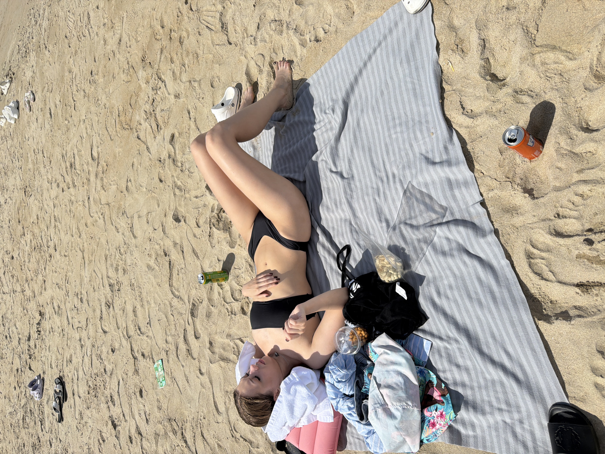 A woman is lying on a gray and white striped beach towel on a sandy beach. She is wearing a black bikini and has a white towel draped over her head and shoulders. A pink pillow is under her head. To her left, on the towel, are various items of clothing, including a floral-patterned shirt, a black top, and a clear plastic bag containing snacks. A can of soda is on the sand to her right. Further in the distance, several pairs of sandals and flip-flops are scattered on the sand, along with a green and white wrapper. Another orange soda can is in the foreground to the right. The scene appears to be Da Nang, Vietnam, as indicated by the location.
