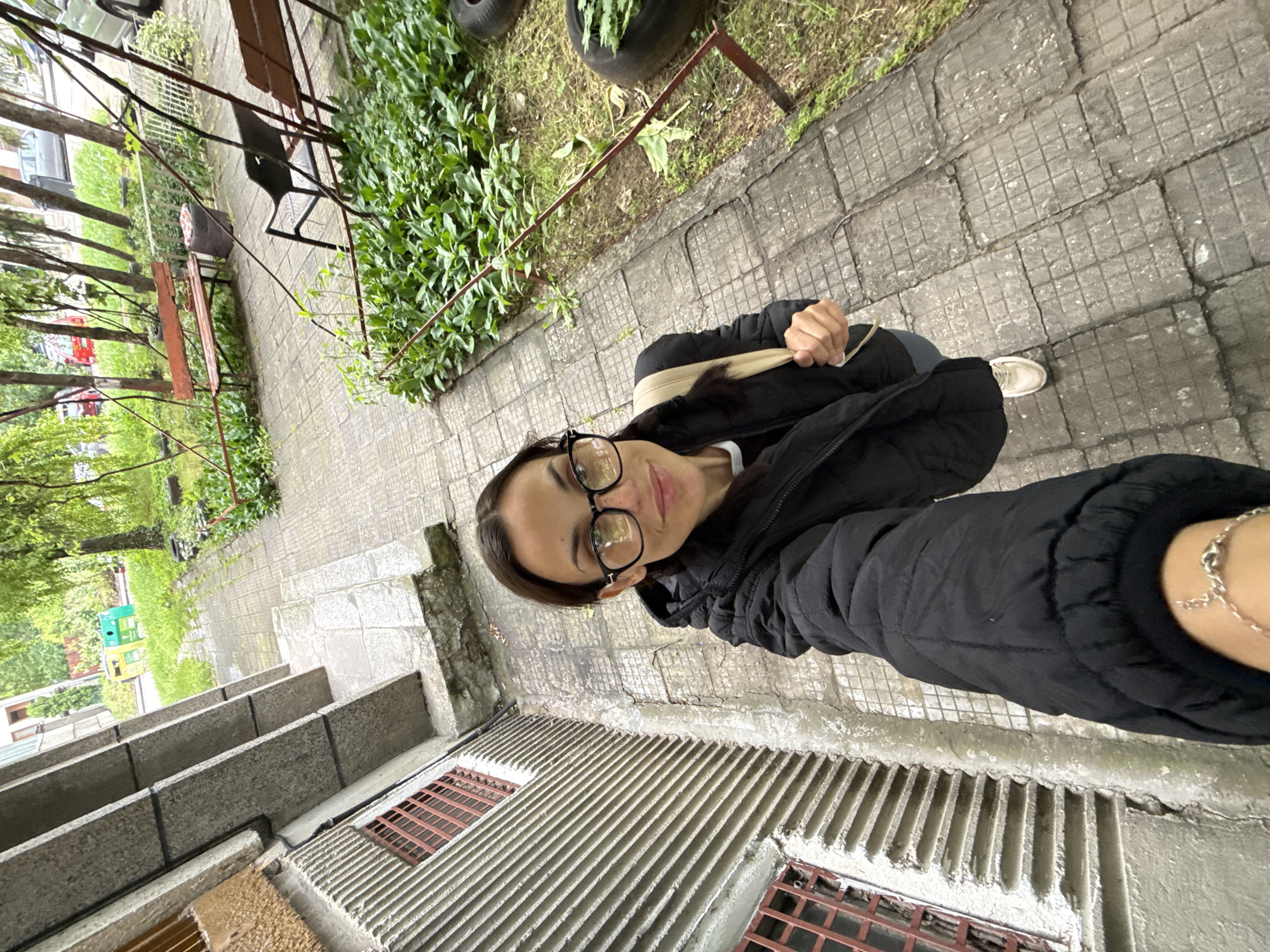 A young woman is taking a selfie outdoors. She wears glasses and a black puffer jacket. She is standing on a concrete surface near a building with metal grating. In the background, there is greenery, trees, and park benches. It is a sunny day. The location is likely in Pleven, Bulgaria.