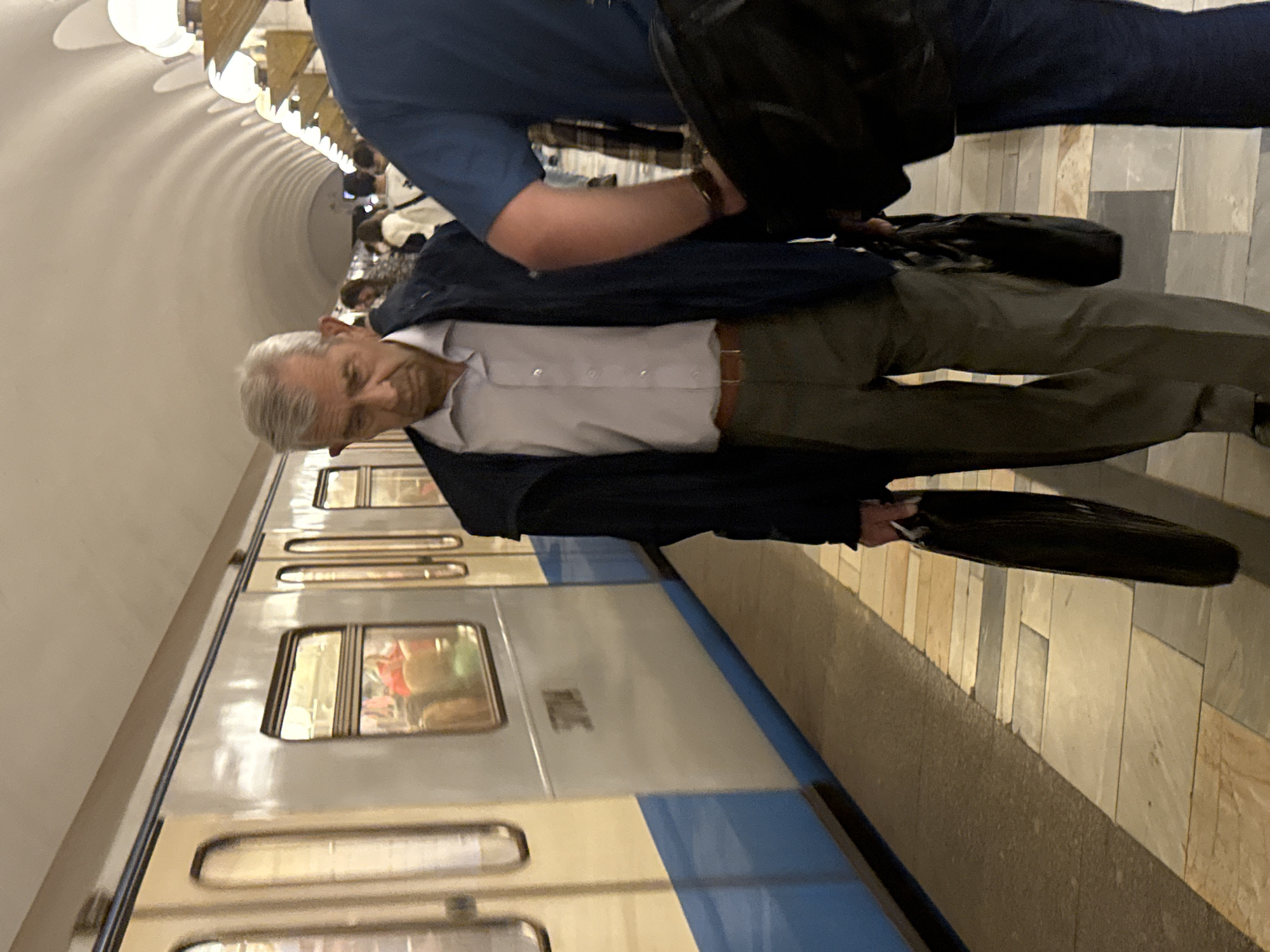An older man stands on a subway platform near a train in Moscow, Russia. He is wearing a light-colored button-down shirt, dark pants, and a dark jacket, and he carries a black briefcase. Another person stands next to him, wearing a blue shirt and dark pants, also holding a black bag. The train is blue and white, and the platform is tiled. The ceiling of the subway station is arched and lit with lights.