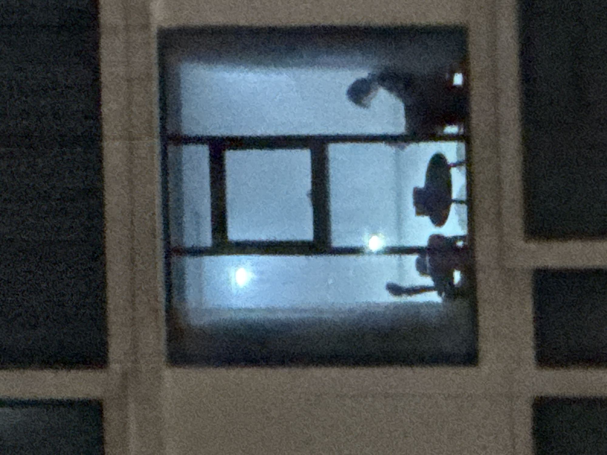 A nighttime view through a window with a grid-like frame. Inside the window, two indistinct figures are visible along with a light source and some furniture, possibly a lamp. The location is unknown.