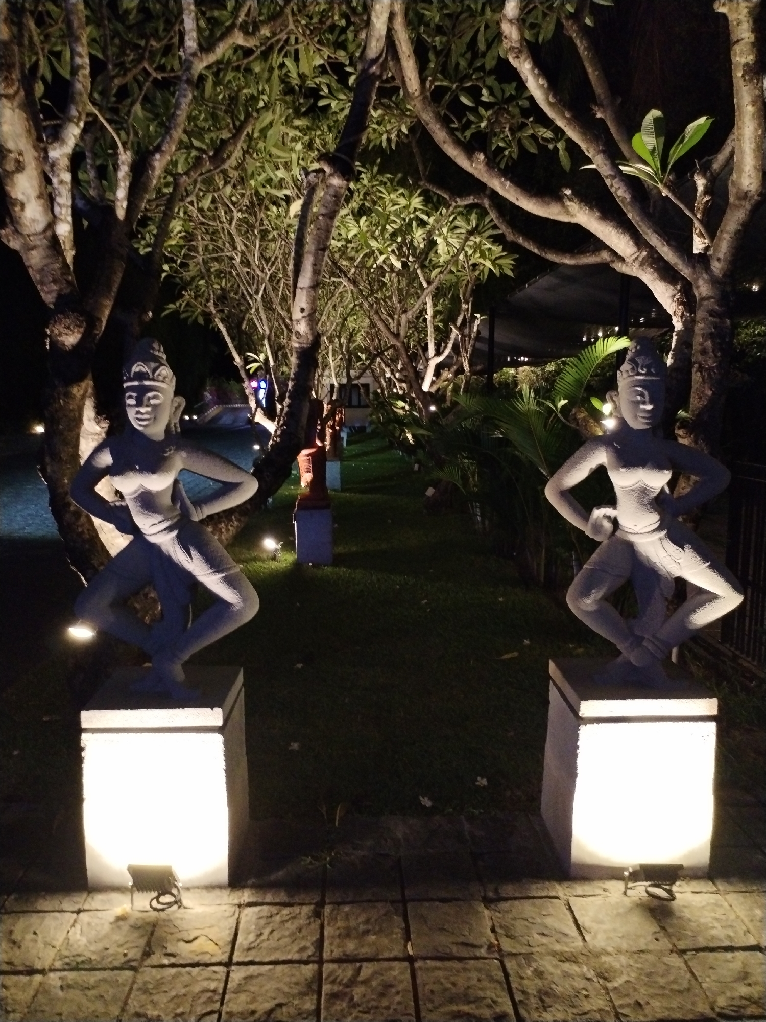 The scene is an outdoor night setting in Bac Nha Trang Ward, Vietnam. Two light-gray stone statues of figures in a dancing pose stand on square, illuminated pedestals on a paved stone surface. Between the statues, a grass lawn extends into the background with additional statues placed along it. Trees surround the area, some appearing to have bare branches, others with green leaves, and they are lit by upward-facing spotlights. Further in the background, building elements are discernible.