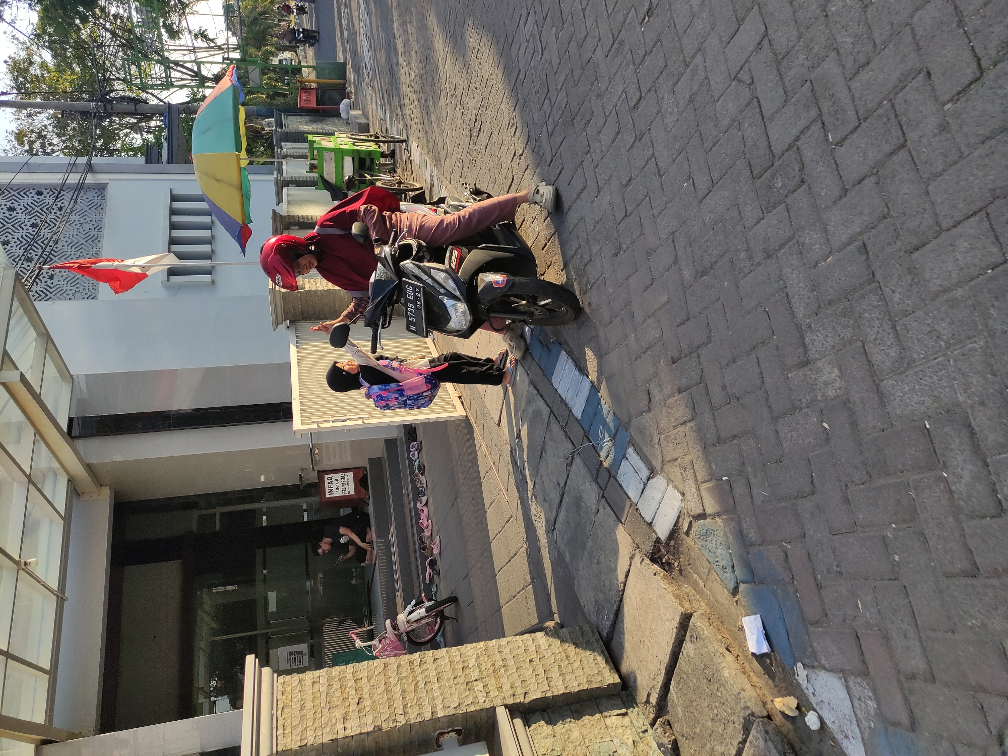 In an outdoor scene, a woman wearing a red helmet and a maroon hijab sits on a motorcycle, facing right. She is dressed in a maroon hijab and a maroon abaya and light brown pants. She is raising her right hand in a wave or gesture. Beside her, a child wearing a black hijab, a black and white patterned backpack, and dark pants stands on the left, also facing right. The child's left hand is extended toward the woman. The motorcycle has a license plate that reads "N 5739 EDG". In the background, a building with a glass entrance and a white patterned wall stands on the left. Several pairs of slippers are arranged on the ground in front of the building. A white bicycle with a pink basket is parked to the left of the motorcycle. A red and white flag flies from a pole on the building. A colorful umbrella is set up in the middle ground, behind the motorcycle. The ground is paved with brick-like stones, and a gray sidewalk with white and blue lines runs along the edge of the brick pavement. Trees and a utility pole are visible in the background. The lighting suggests it is daytime, with shadows cast on the ground.