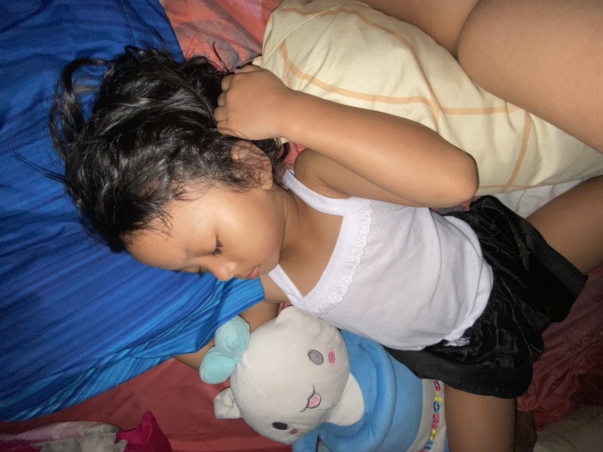 A young child is sleeping soundly in bed, nestled with a stuffed animal. The child, with dark hair, is wearing a white tank top and black shorts. A light-colored pillow with orange stripes is partially visible under their head. The bedding appears to be a mix of colors, including blue, red, and pink. The scene suggests a cozy and peaceful moment, likely during the night given the dim lighting. The child is in a relaxed sleeping pose, with one arm resting on the pillow and the other seemingly holding the stuffed toy. The stuffed animal is blue and white with embroidered facial features, including pink cheeks and a smiling mouth. The overall impression is one of comfort and security. No visible text is present in the image. The location context provided is Gelam, Indonesia.