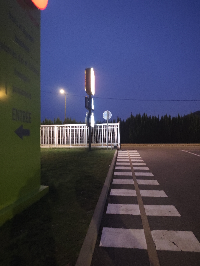 A crosswalk stretches alongside a grassy median, bordered by a low, white fence. Behind the fence stand two tall signs, one glowing with an elongated, rounded rectangular shape and the other circular. A street lamp is visible on the left. Coniferous trees fill the background beneath a dark blue sky. A green building with an arrow pointing left and the word "ENTREE" is on the left edge of the image.