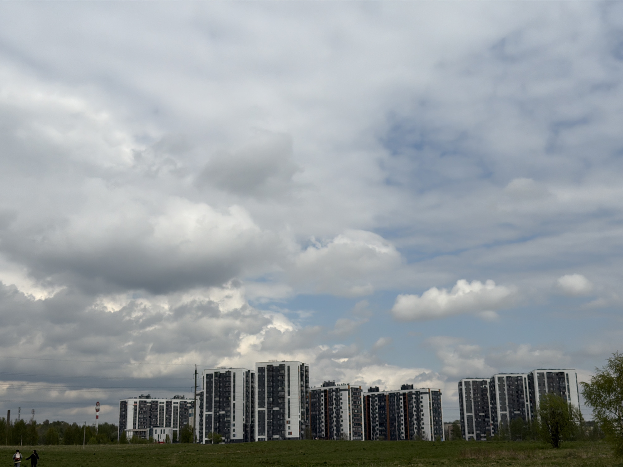 The image shows a low-angle view of the apartment complex in Yanino-1, Russia. The complex consists of multiple high-rise apartment buildings with a white and grey color scheme. A field of green grass extends from the bottom of the frame towards the buildings. In the background, a few trees are present to the left and right. In the lower left corner, two figures walk along a path. Above the buildings, the sky is filled with a mix of dark grey and light white clouds. Some power lines are visible in the distance.