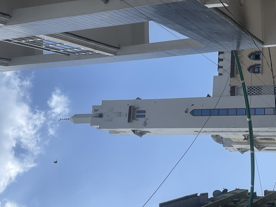The image displays an upward perspective of the Malindi Mosque's minaret in Stone Town, Zanzibar City, Tanzania, under a clear blue sky with scattered white cumulus clouds. The minaret, a tall, slender, white concrete structure, occupies the central frame. Its apex is a pointed spire crowned with a metallic crescent moon and star finial. Below the spire, four dark loudspeakers are affixed, and multiple rectangular, blue-tinted glass windows extend vertically down one side. Weathering and minor paint chipping are evident on the minaret's white facade. A single dark-colored bird is captured in flight in the upper-left quadrant of the sky. To the right, a portion of a modern, multi-story building with a light grey tiled exterior and structural columns is visible. To the left, an older building, partially obscured, shows elements like air conditioning units and satellite dishes. Several dark utility cables and bundled green cables intersect the view across the lower and middle sections of the image. Structures comprising the mosque's main body and an adjacent yellow-painted building with arched windows are visible in the background. No human subjects are present.