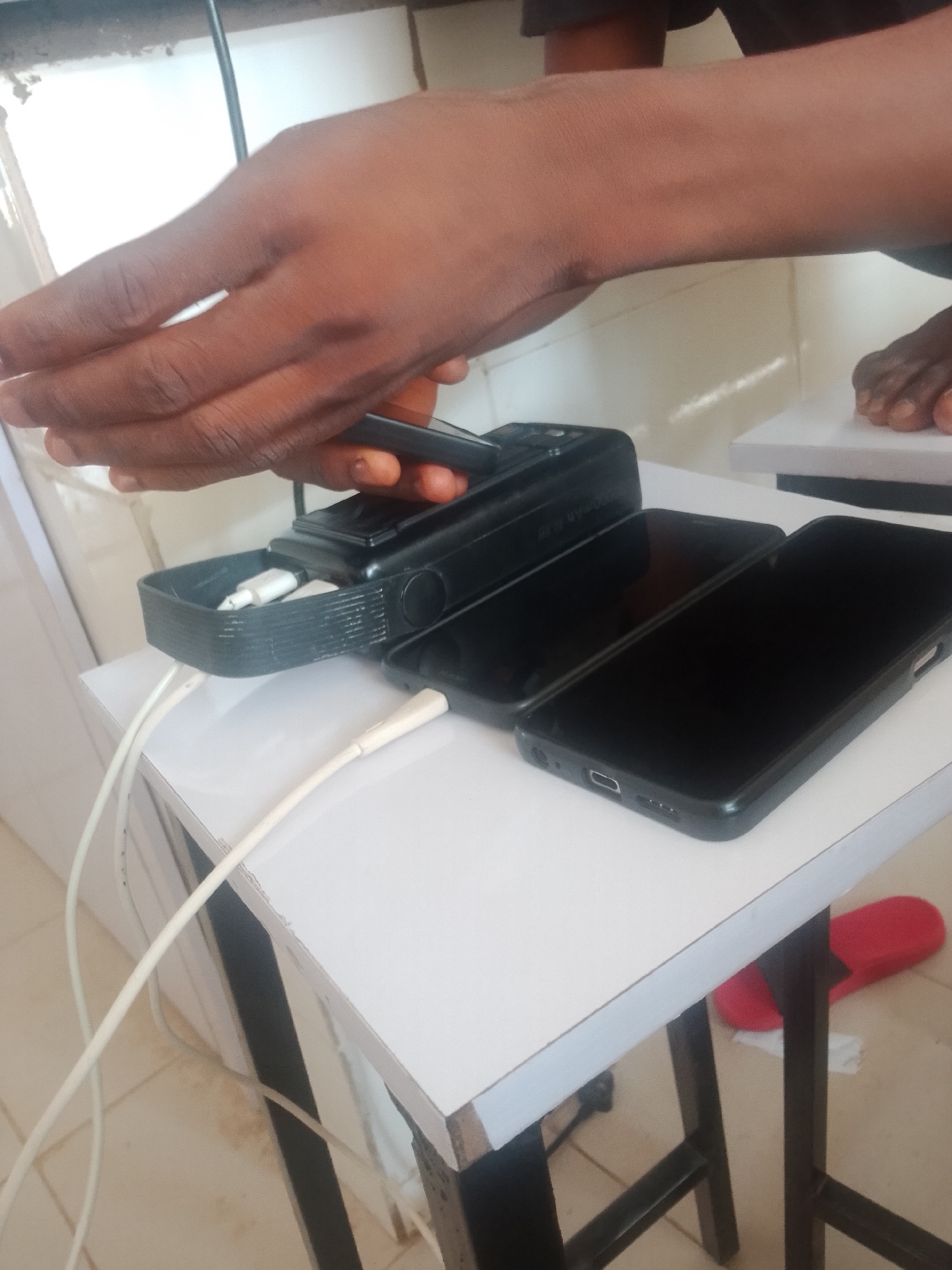 The image shows a person's hands holding a cell phone, with two other cell phones connected to a black charging device on a white table. The charging device has a small compartment holding two white charging cables. The scene is indoors, with a tiled floor visible beneath the table. There is a red slipper on the floor to the right of the table.