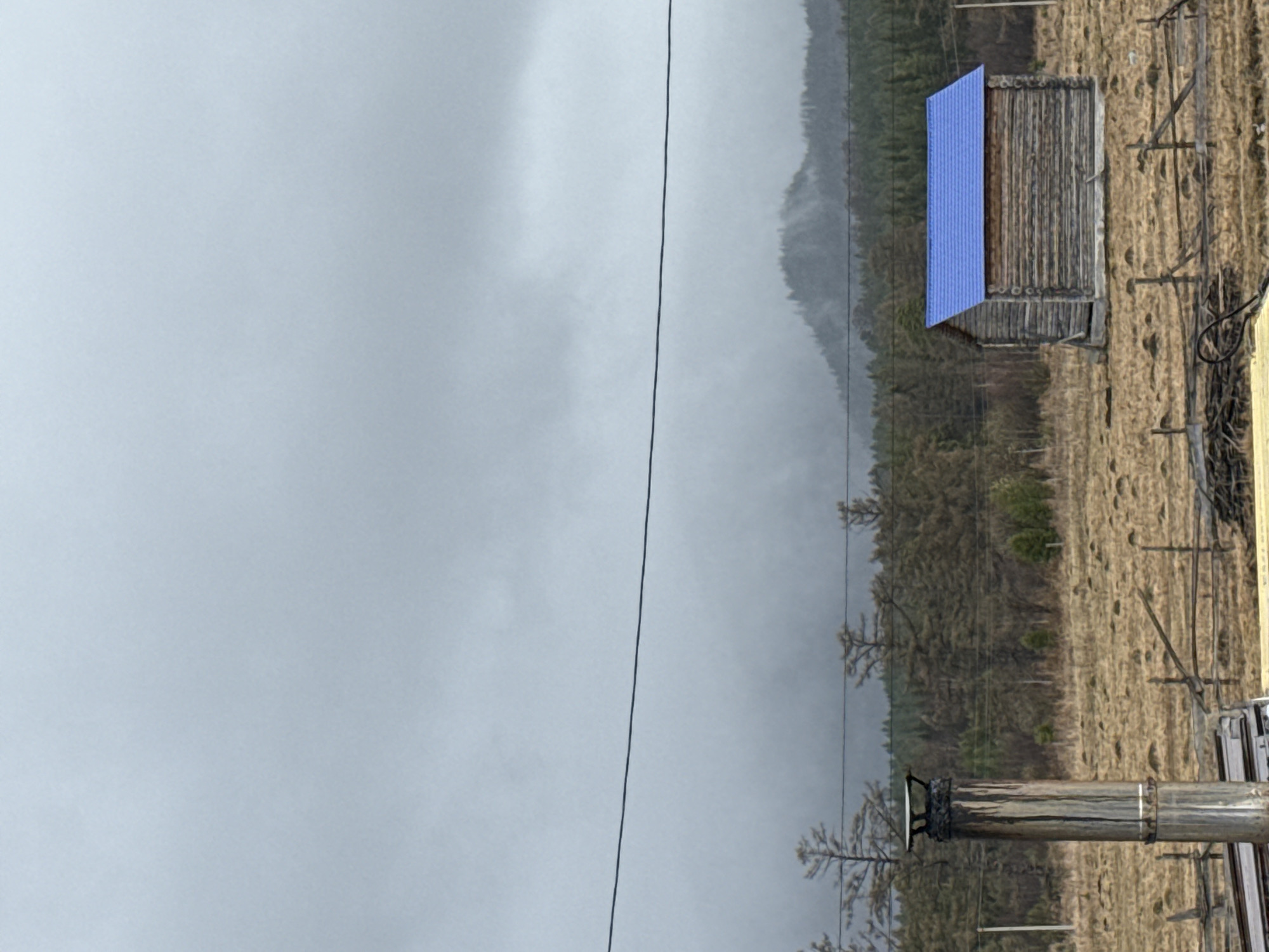 The image shows a landscape in Tyulyuk, Russia. The left side is dominated by a gray, overcast sky. A power line bisects the image vertically. In the middle ground, there is a dense forest transitioning to a meadow. A small wooden structure with a blue roof stands out in the meadow. A metal chimney stack is in the foreground. The overall scene suggests a rural or remote location in a mountainous region under a heavy sky.