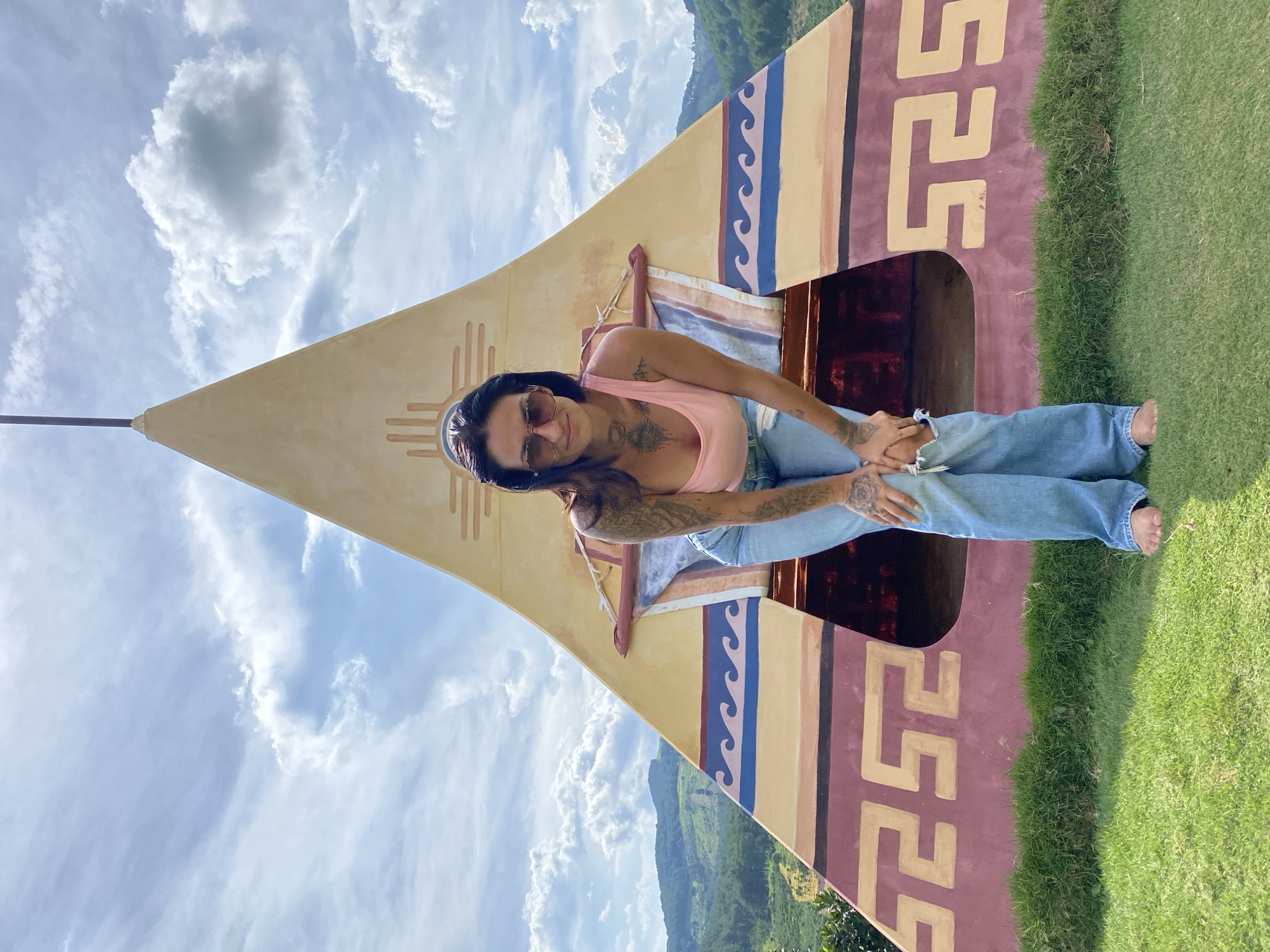A woman with tattoos and sunglasses is sitting on a colorful, triangular object with indigenous-inspired patterns. She is wearing a pink tank top and light blue jeans. The object appears to be a decorative or artistic installation, possibly a prop. The background shows a bright blue sky with fluffy white clouds and a lush green hillside. The image is taken from a low angle, emphasizing the sky and the structure. The lighting suggests it is daytime, and the weather is clear and pleasant. The overall mood is casual and relaxed. The image is oriented vertically, with the subject positioned in the center and to the left. The grass in the foreground is neatly trimmed.