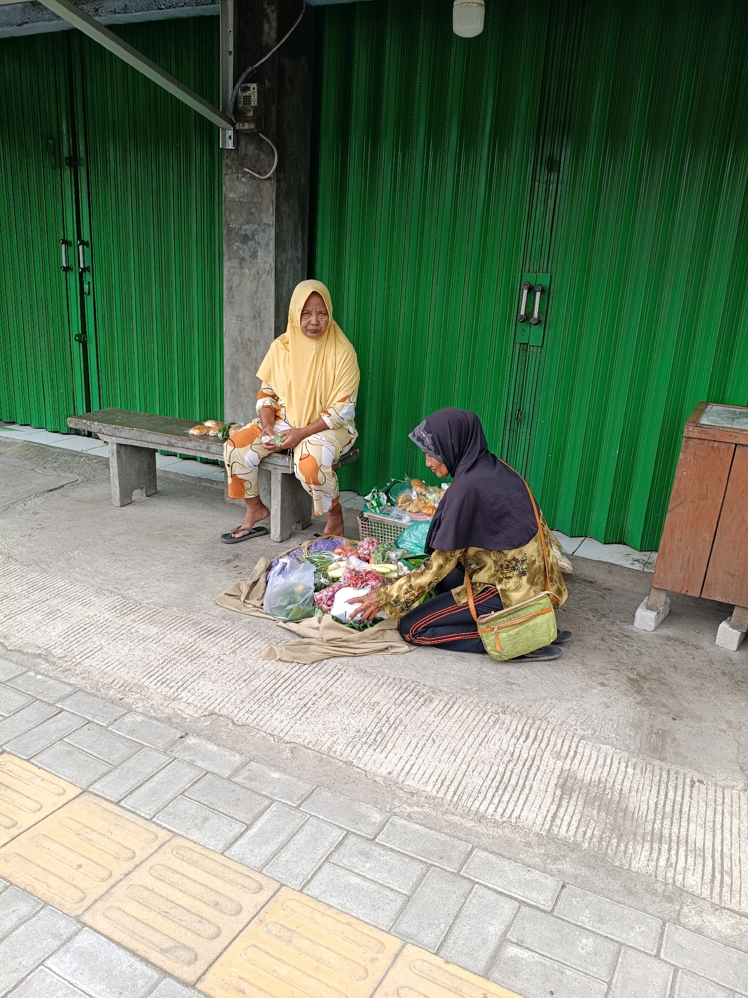 Two women are selling produce on the sidewalk in front of a green shuttered building in Sewon, Indonesia. The woman on the left is sitting on a concrete bench, wearing a yellow headscarf and patterned pants, and appears to be holding a small item in her hands. In front of her on the bench are some packaged food items. The woman on the right is kneeling on the ground, wearing a black headscarf, a patterned top, and black pants. She is arranging vegetables and other produce spread out on a tan cloth on the ground. A basket of produce is next to her, and she has a green shoulder bag. The sidewalk in front of them is made of concrete with textured lines and yellow tactile paving stones. A wooden structure with two stone blocks supporting it is to the right of the scene. The background features a tall, green, corrugated metal shuttered building.