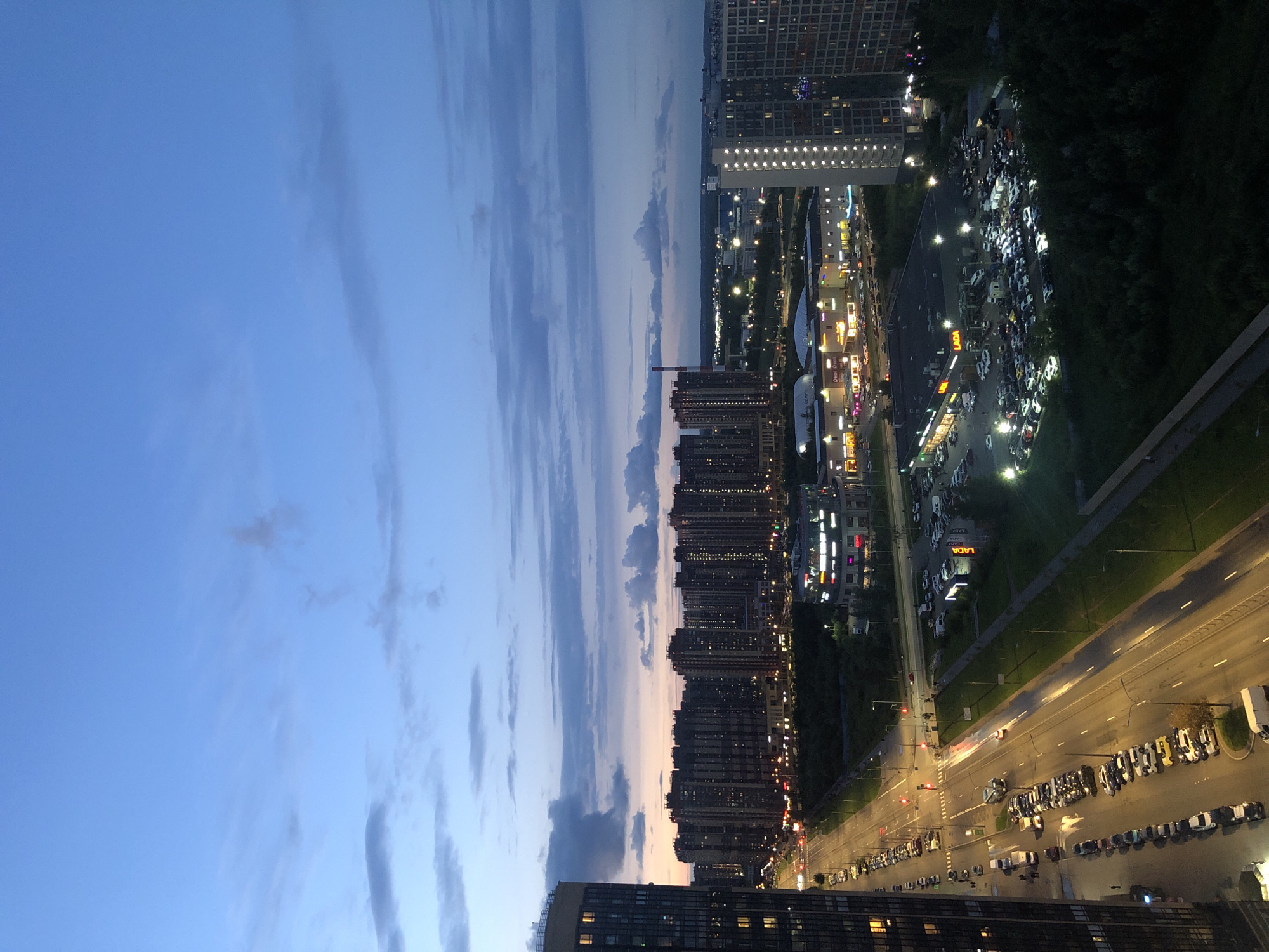 The image is an aerial view of a city street at dusk. A multi-lane road runs diagonally across the frame, lined with parked cars and illuminated by streetlights. Several modern residential buildings with many windows are visible on the left side of the road, one of which is in the foreground. Across the street, a large shopping center with illuminated signs and a parking lot filled with cars is situated. Beyond that, a series of taller, dark buildings form a cityscape, with some lights visible in their windows. The sky above is a deep blue, with scattered clouds. In the distance, more buildings and lights indicate a sprawling urban area.