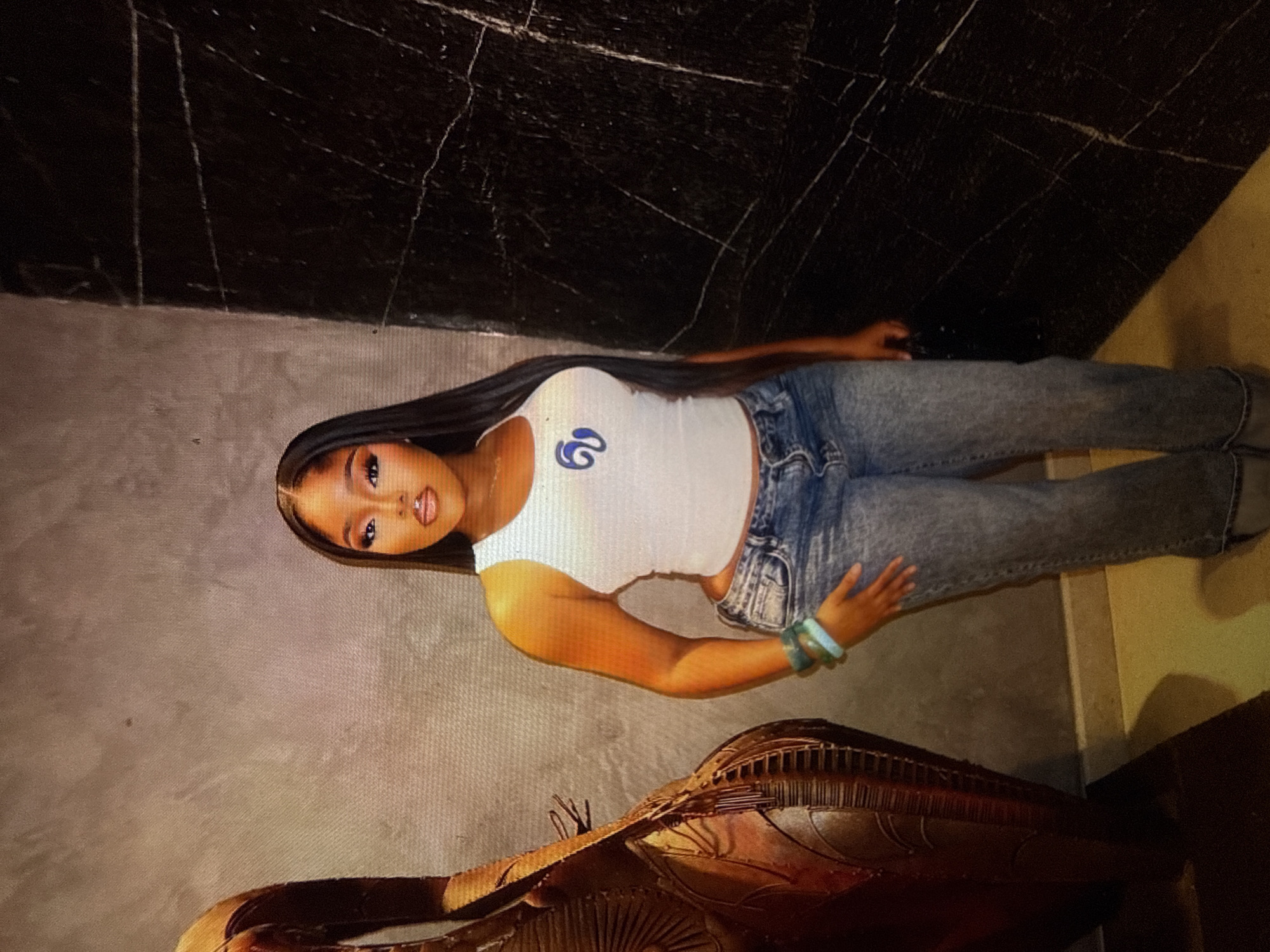 A young woman stands in what appears to be an indoor setting with textured walls. She has long, dark hair, is wearing a white tank top with a blue graphic, and light-wash jeans. She is also wearing bracelets on her left wrist and appears to be holding a dark clutch bag. To her left is a tall, ornate decorative object with intricate carvings. The wall behind her is a mottled grey on the left and a dark, veined material on the right. The floor is a light yellow color. The scene does not contain any discernible features that would indicate its location within the city of Abuja, Nigeria.