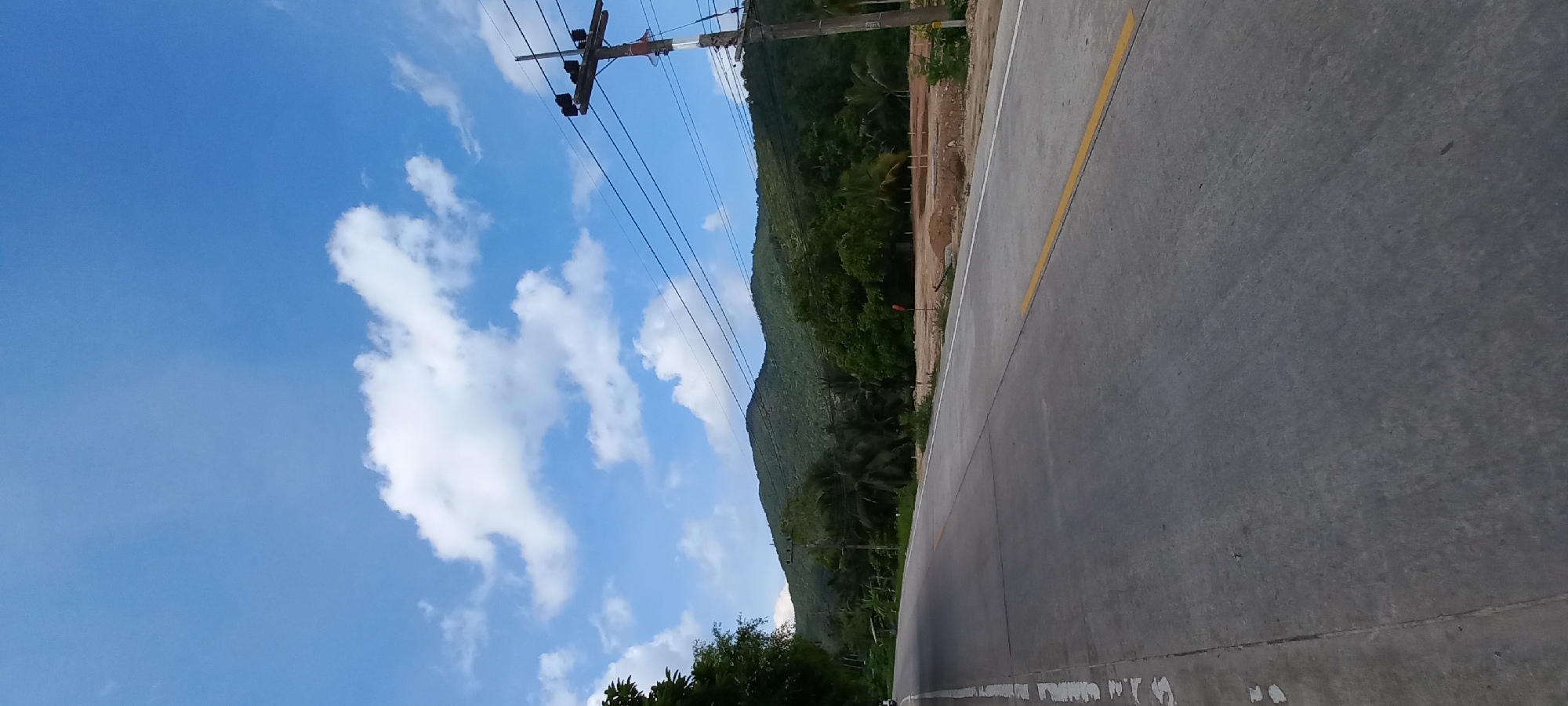 The image shows a road in Baan Chaloklum, Thailand. The road is concrete with a yellow line marking the center. A mountain covered in dense green vegetation rises on one side of the road. Utility poles with power lines run along the edge of the road. The sky is blue with scattered white clouds. Part of what looks like a construction or excavation site is also visible between the road and the mountain vegetation.
