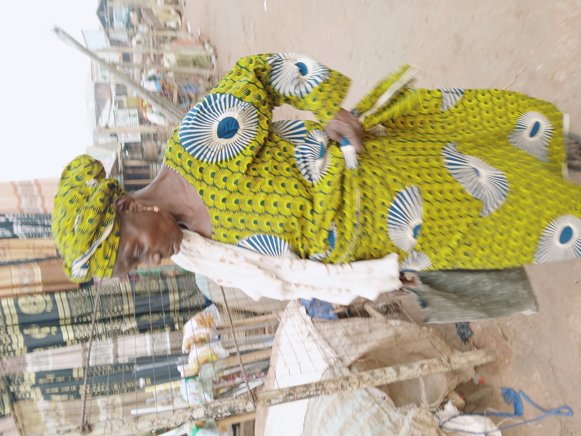 In Ankpa, Nigeria, an older woman with brown skin is sitting on a low seat. She is wearing a headwrap and a patterned dress with a yellow background and large blue and white circular designs. A white garment is draped over her shoulders. She has a gold earring. The background suggests a market setting with items displayed for sale and rudimentary structures. The ground appears to be dirt or a similar material.