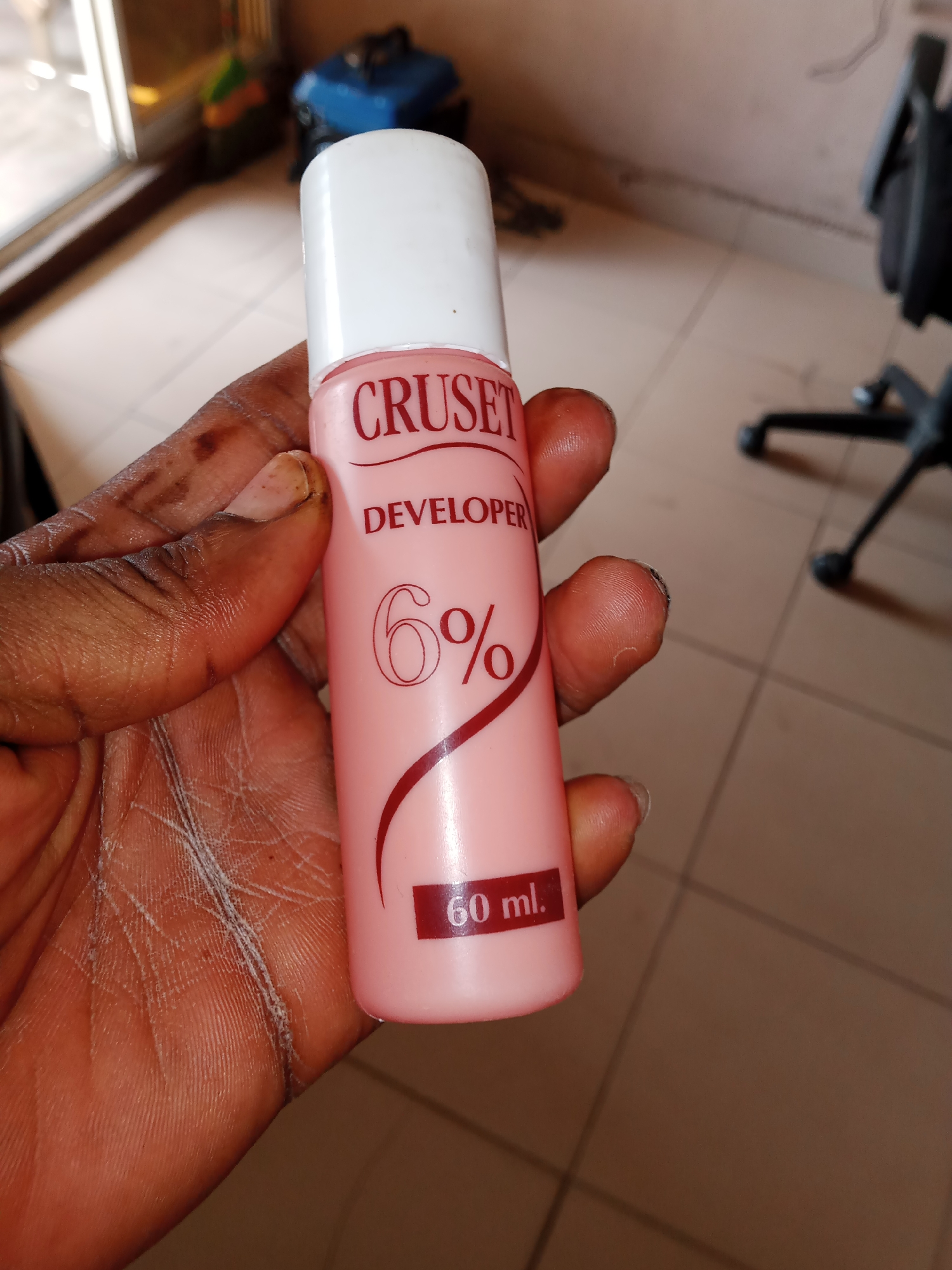 A close-up shot focuses on a pink bottle of "CRUSET DEVELOPER 6% 60 ml" being held by a person's hand. The bottle has a white cap and red text. The hand appears to be of African descent with visible skin texture and fingernails. In the blurred background, what appears to be a portion of an office chair is visible, along with a tiled floor and a window with light coming through. The scene is indoors. This image was captured in Jalingo, Nigeria.