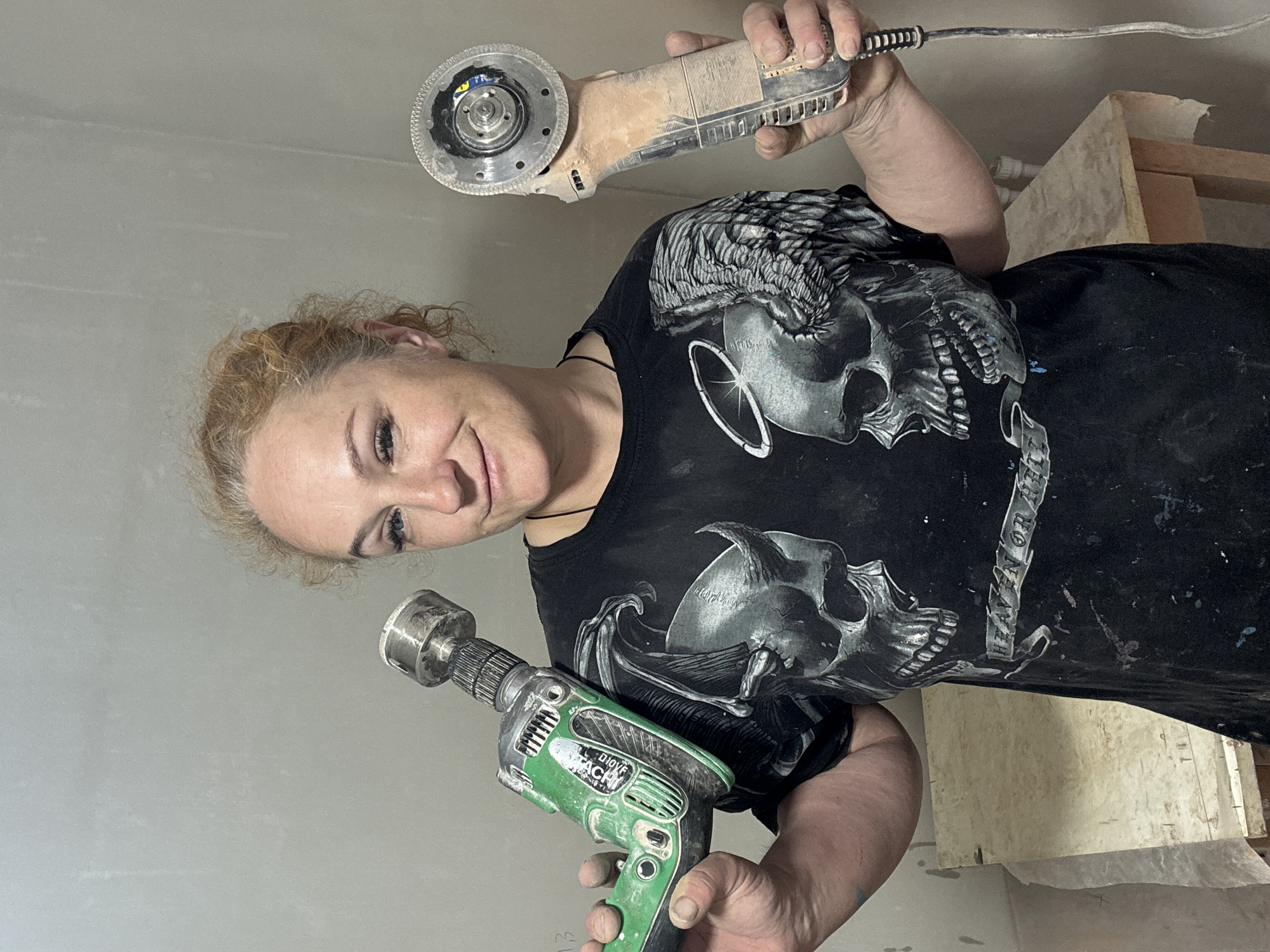 A woman is holding a drill in one hand and a grinder in the other. She is wearing a black t-shirt with a skull graphic. The background appears to be an unfinished room with light-colored walls. It is impossible to ascertain the location in the city of Chaikovsky, Russia.