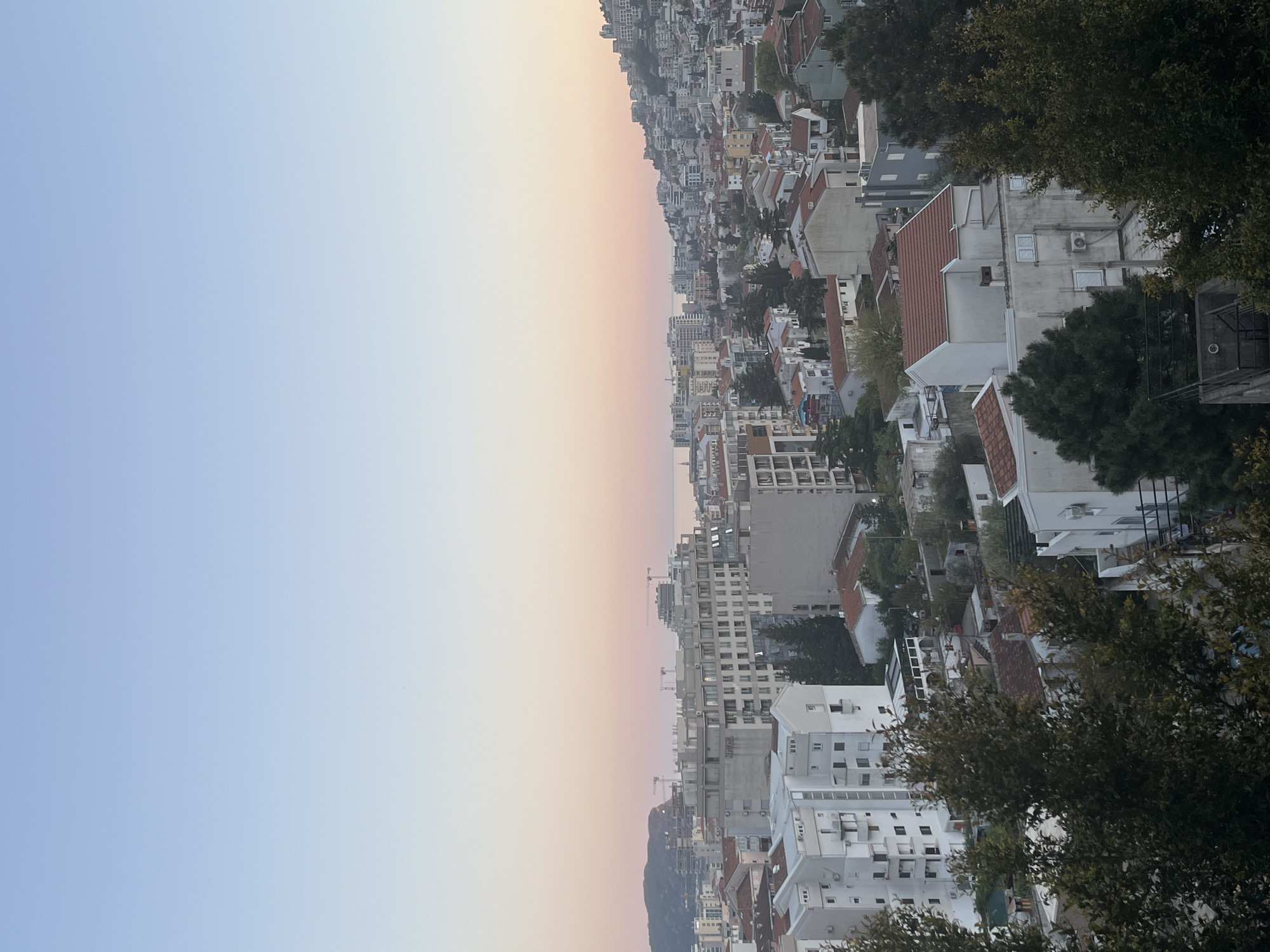 The image shows a high-angle view of a cityscape at dusk. The sky fades from blue to orange, indicative of sunset or sunrise. The city itself is densely packed with buildings of varying heights and architectural styles, mostly residential, with a mix of white, red-tiled roofs, and other light-colored structures. Trees are interspersed throughout the cityscape. Given the coastal location and building style it could be the city of Budva, Montenegro. There is no sign of people, objects, actions, or interactions.