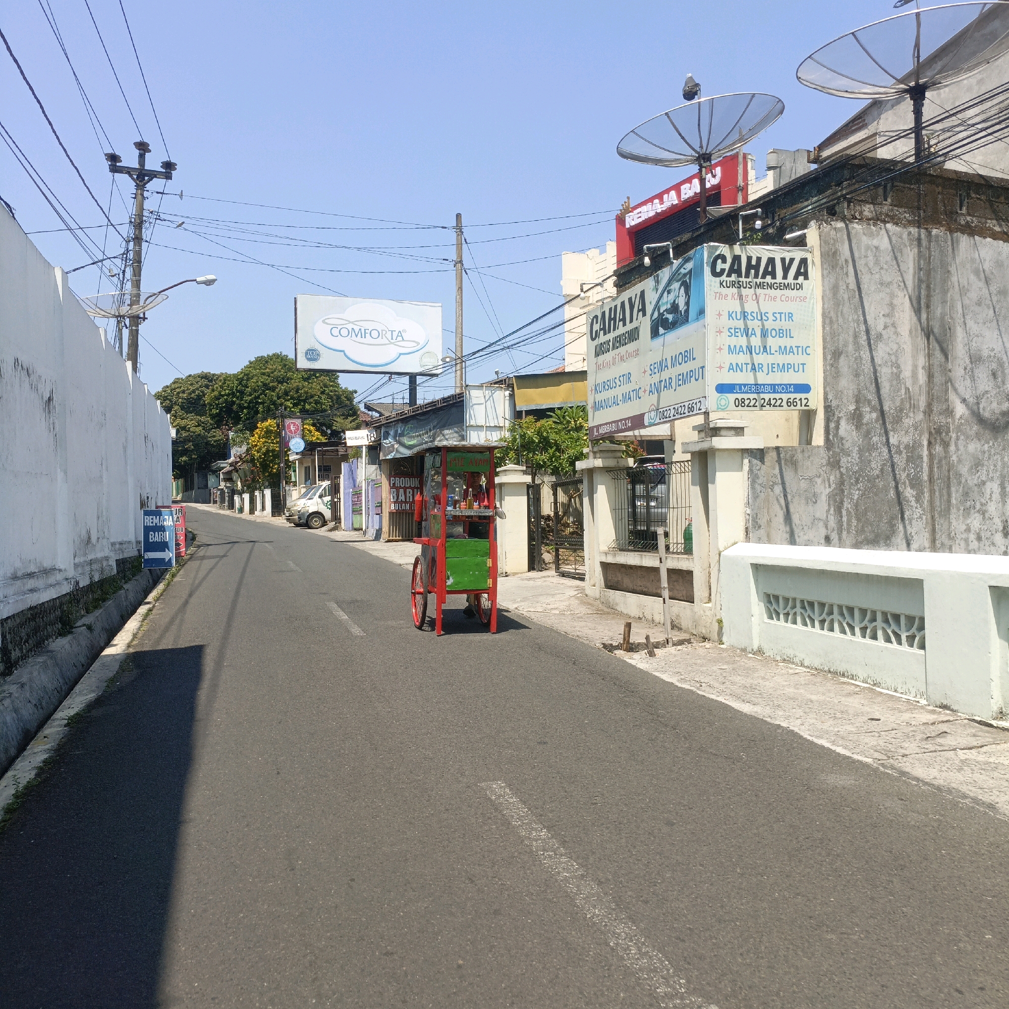 A street scene in Salatiga, Indonesia, is captured under a clear blue sky on a sunny day. The image focuses on a street with an asphalt surface, marked with a white dashed line. On the left, a tall, textured white wall runs along the side of the road, with a narrow drainage ditch at its base. A blue and red sign with white text and an arrow pointing to the right is affixed to the wall.
In the center of the street, a colorful food cart with a red frame and green compartments is stationary. The cart has two large red spoked wheels and a red canopy. A person is visible behind the counter of the cart.
Buildings line the right side of the street. A multi-story building with a concrete facade is prominent, featuring two large satellite dishes mounted on its roof. A red sign with white text, "REMAJA BAJ," is visible on this building. Below this, a large vertical banner advertises "CAHAYA KURSUS MENGEMUDI" (Cahaya Driving Course) and lists services such as "KURSUS STIR" (steering course), "SEWA MOBIL" (car rental), "MANUAL-MATIC," and "ANTAR JEMPUT" (pick-up and drop-off). A phone number, "0822 2422 6612," is also displayed. Further down the street, more buildings and trees can be seen. A white van is parked on the right side of the road. Electrical poles and wires are visible throughout the scene. The overall environment appears to be a moderately developed urban or suburban area.