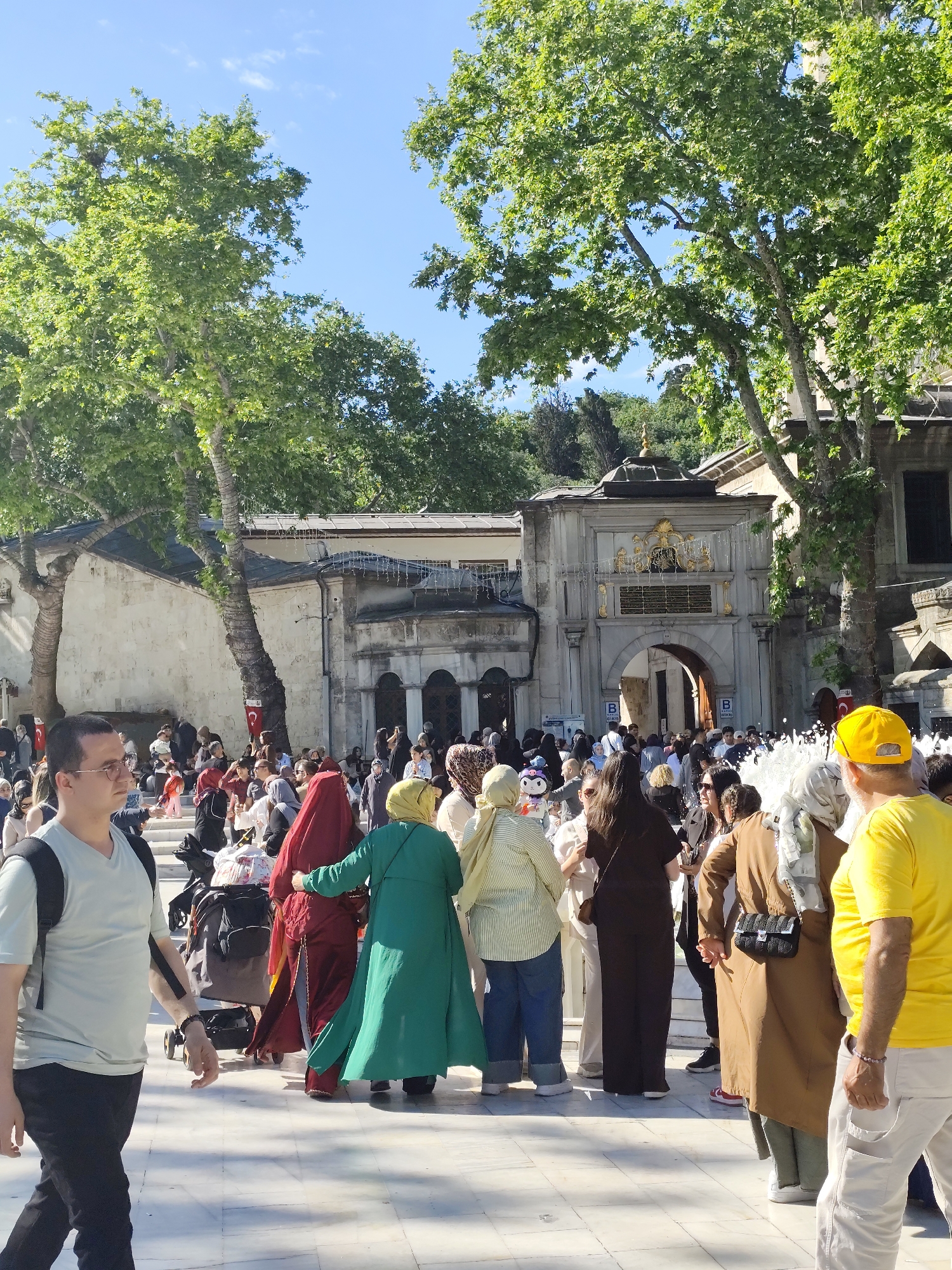 A daytime outdoor scene shows a crowded public square in Eyüpsultan, Turkey. A diverse group of people, including women wearing hijabs of various colors and styles, populate the area in front of an ornate, arched gateway, which marks the entrance to a historic complex. The gateway is embellished with gilded details and flanked by stone structures with arched windows. Lush green trees frame the architectural elements and provide shade. The sky is a clear, pale blue. In the foreground, a man wearing a light-colored shirt and black pants walks to the left. A man wearing a yellow shirt and yellow cap is partially visible at the right of the frame. Numerous other individuals, including those pushing strollers, are seen interacting and moving through the square. The ground is paved with light-colored stone tiles.