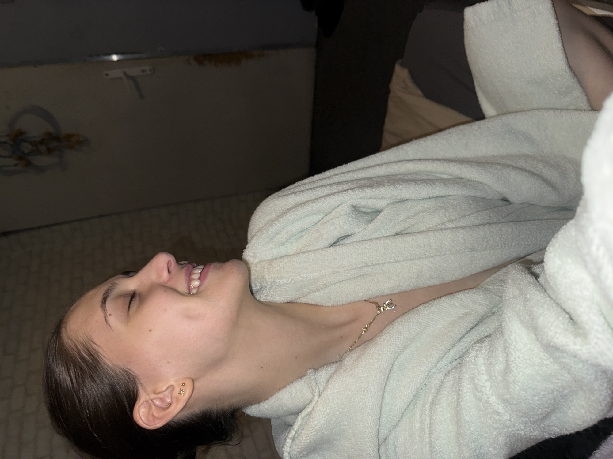 A young woman with dark hair is lying on a bed, wearing a light-colored terry cloth robe and a delicate gold necklace. She is smiling slightly with her eyes closed, facing upwards and to the left. The background features a wall with faint decorative elements and a dark object, possibly clothing. The image was not taken in Sofia, Bulgaria, as no elements in the photo indicate that location.
