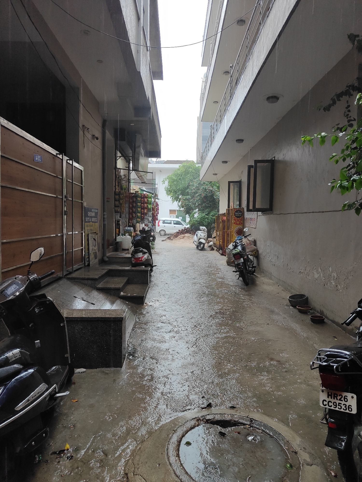 The image depicts a narrow, wet alleyway in Gurgaon, India, under a steady rainfall. The wet asphalt ground reflects the overcast sky and the surrounding buildings. On the left, a brown wooden gate with a blue sign indicating "A-512" is prominent. Adjacent to it is a building with a concrete facade and a store selling various goods displayed on shelves. Several scooters are parked in the alleyway, including a dark colored scooter in the foreground on the left and a black motorcycle with a license plate reading "HR26 CC9536" on the right. In the background, a white car is visible, and there are stacks of materials, possibly construction-related, on the right side. The buildings on both sides of the alley are multi-story structures with balconies and windows. The overall scene conveys a sense of a residential or commercial street in the midst of a downpour.