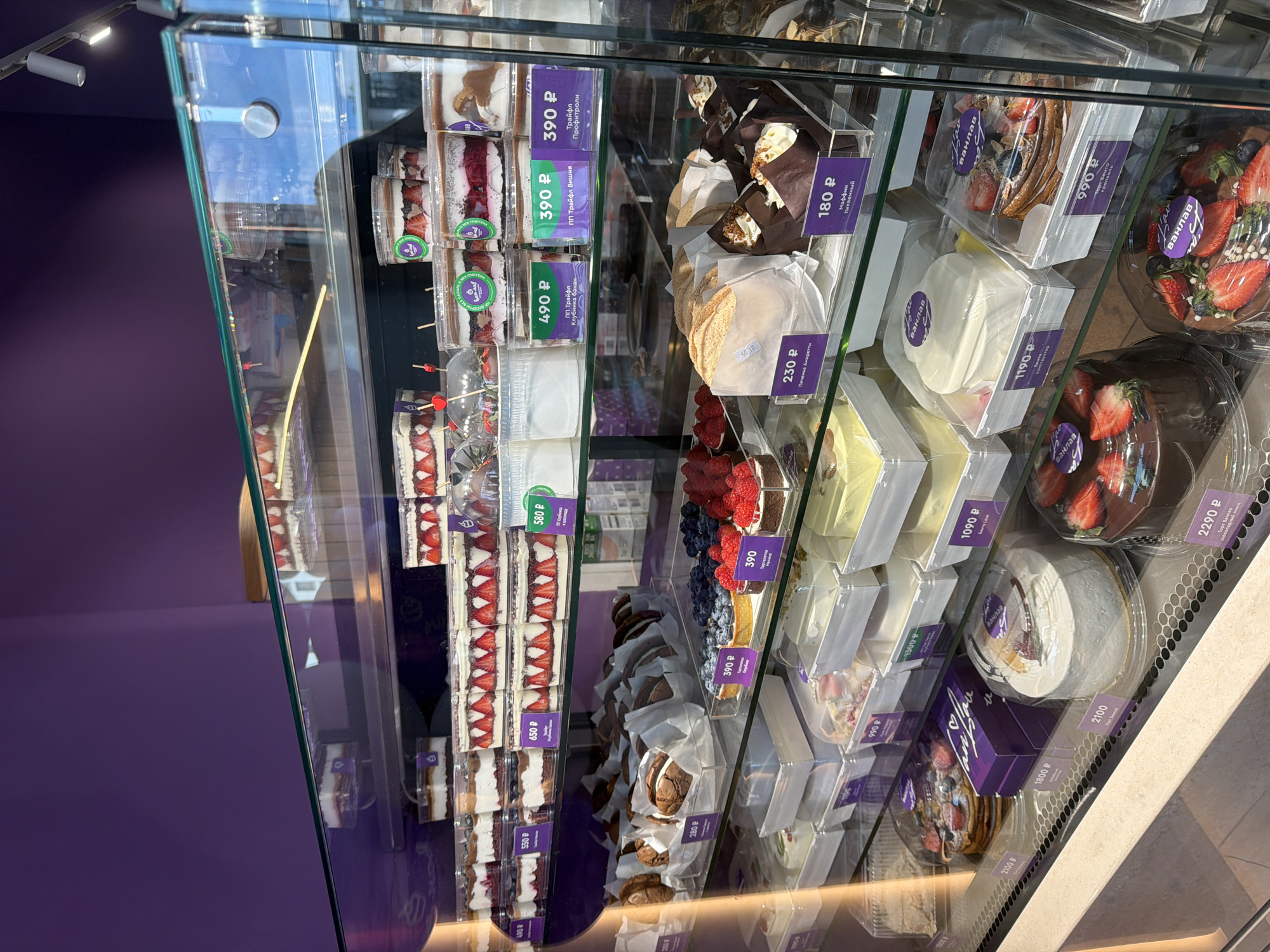 The image displays a brightly lit glass display case filled with an assortment of desserts for sale in a bakery or dessert shop. The interior features a distinctive purple wall, which is reflected in the glass, along with overhead ceiling lights.
Numerous individual desserts are neatly arranged on multiple shelves within the case. These include various types of trifles, presented in clear plastic cups, often layered with cake, cream, and fruits like strawberries, bananas, and cherries. Other sweet treats visible are muffins, cookies (specifically "Печенье Амаретти" - Amaretti Cookies), and tarts abundantly topped with fresh berries such as raspberries and blueberries. Larger cakes, some branded "Ванлав" (Vanlav), are also present in clear plastic packaging, including "Бенто Love" and "Торт Бенто Lav," suggesting individual or small-sized cakes.
Each dessert has a price tag, primarily purple or green, indicating prices in Russian Rubles (P). Notable visible text includes:
* "ПП Трайфл Клубника банан" (PP Trifle Strawberry Banana) for 490 P.
* "ПП Трайфл Вишня" (PP Trifle Cherry) for 390 P.
* "Трайфл Профитроли" (Trifle Profiteroles) for 390 P.
* "Маффин тыквенный" (Pumpkin Muffin) for 180 P.
* "Печенье Амаретти" (Amaretti Cookies) for 230 P.
* "Бенто Love" for 1090 P.
* "Ванлав" branding on several cakes, with prices up to 2290 P for a "Торт Ванлав адный мини."
The display is well-organized, showcasing a variety of textures and colors from the different pastries and fresh fruits. No people are visible, emphasizing the selection of available items.