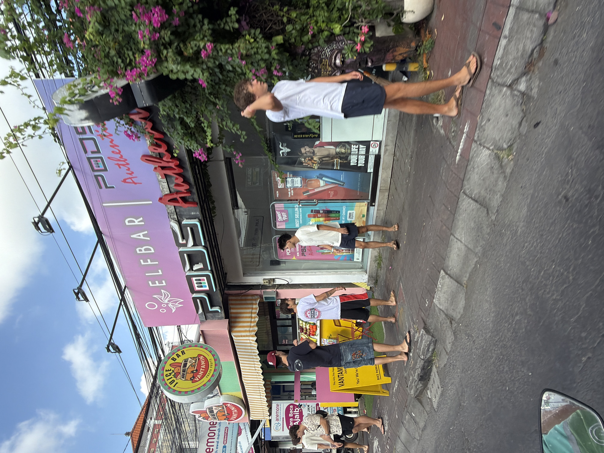 In North Kuta, Indonesia, on a street lined with shops and businesses, a group of young men stand on the sidewalk in front of a vape shop called "PODS Authentic". One man in the foreground, wearing a white t-shirt and dark shorts, walks past the shop. Behind him, other men are gathered, some looking at their phones, others talking. The vape shop has large windows displaying various vaping products and promotions. A brightly colored "JUICE BAR" sign is visible to the left of the vape shop, along with other signage for businesses. The street is paved, with a sidewalk and a gutter. The sky above is blue with some clouds, and there is lush green foliage with pink flowers growing on the side of the building. The overall scene depicts a typical urban street environment in a commercial district.