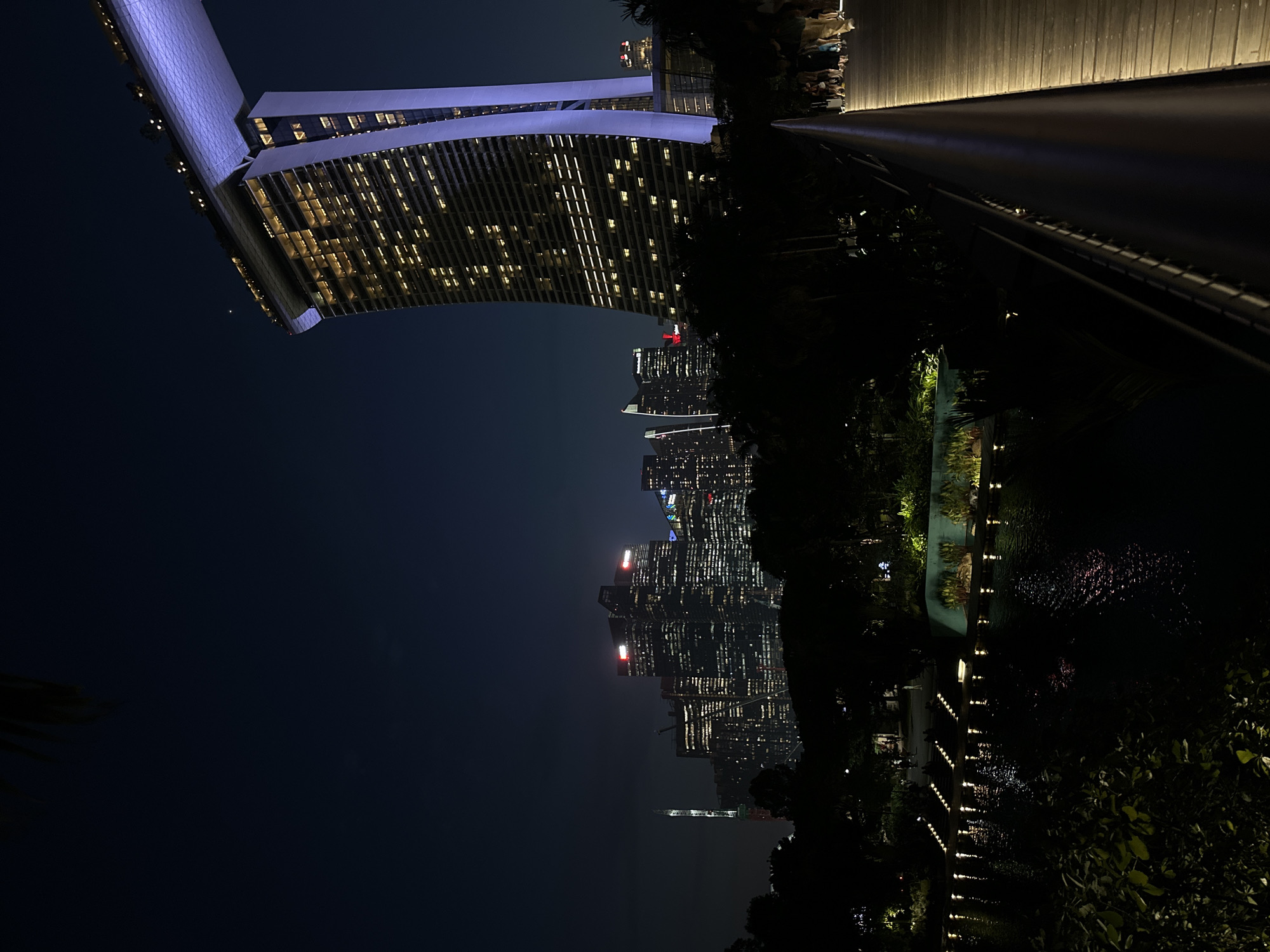 The image depicts a night view of the Marina Bay Sands in Singapore. In the foreground, a wooden walkway with a railing lines the right side of the frame. Beyond the walkway, lush green foliage and trees obscure the ground level. A body of water, reflecting the ambient light, occupies the mid-ground. Rows of illuminated lights are visible along the edge of the water. In the background, the iconic Marina Bay Sands hotel stands tall, its illuminated windows creating a grid pattern on its facade, and its sky-high infinity pool area is bathed in a soft purple light. To the left of the hotel, the Singapore skyline is visible, with numerous skyscrapers illuminated by their own lights, some featuring red accents at their tops. A few people are visible walking on the distant walkway, their figures silhouetted against the light. The overall atmosphere is one of urban nighttime grandeur and serene natural elements.