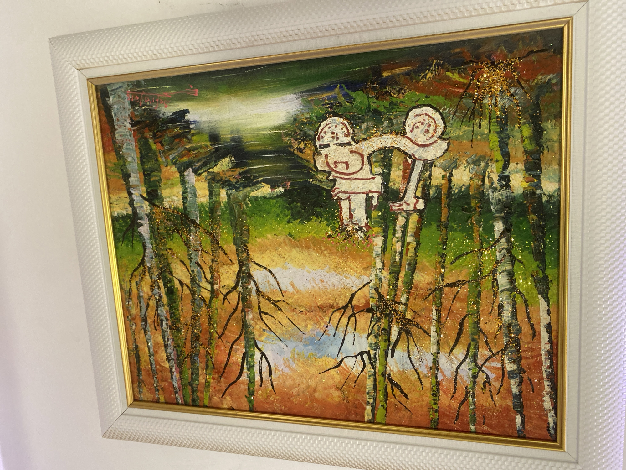 A framed painting hangs on a white wall. The painting depicts a forest scene with birch trees. Two stylized figures, outlined in red, are positioned among the trees, appearing to hold hands. Gold glitter is scattered throughout the painting, particularly on the trees and above them. The painting is framed with a white, textured outer frame and a thin gold inner frame. The background colors are green, brown, orange, and a hint of blue. The style of the painting is naive and abstract.