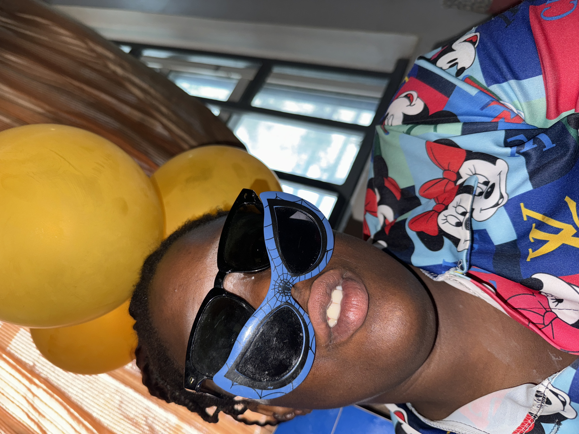 A person with dark skin is wearing sunglasses with a blue frame and spider web design on the lenses. They are also wearing a colorful shirt with a cartoon print of Mickey Mouse. Three yellow balloons are visible in the background. The location appears to be indoors, possibly Sabon Tasha, Nigeria. The image is rotated 90 degrees counterclockwise.