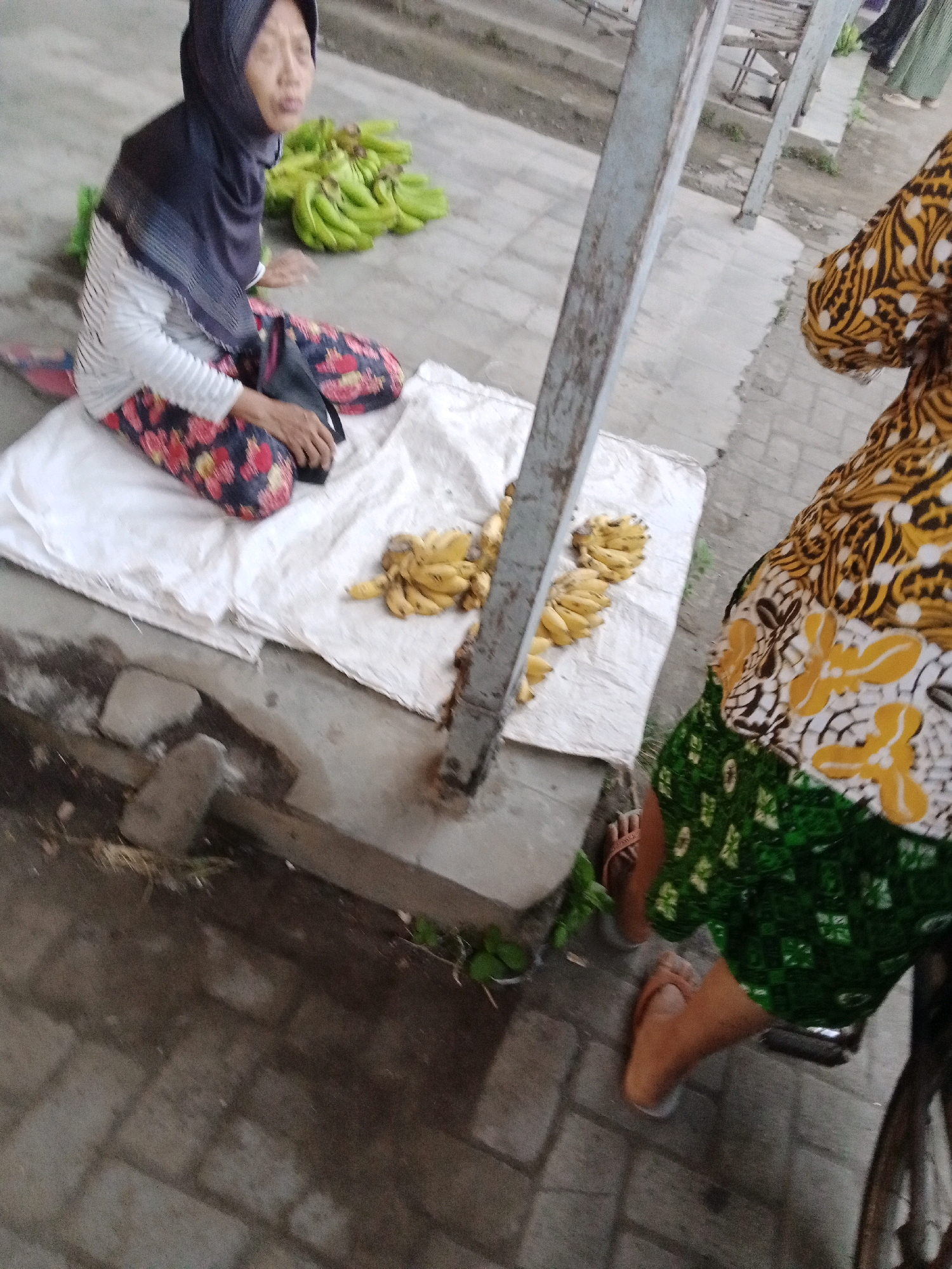 An outdoor scene in Ngronggot, Indonesia, shows two individuals at what appears to be a market stall. The person in the foreground is seated on the ground, wearing a dark hijab, a striped shirt, and patterned pants. They are next to a pile of green bananas and a small bunch of yellow bananas displayed on a white cloth. To the right, another person, partially visible, is wearing a brightly colored, patterned outfit with a green skirt and sandals. Their legs and feet are visible as they stand. The ground is paved with brick-like stones. A metal post stands vertically between the two individuals. The overall impression is of a casual market environment, possibly in the late afternoon or early evening given the lighting.