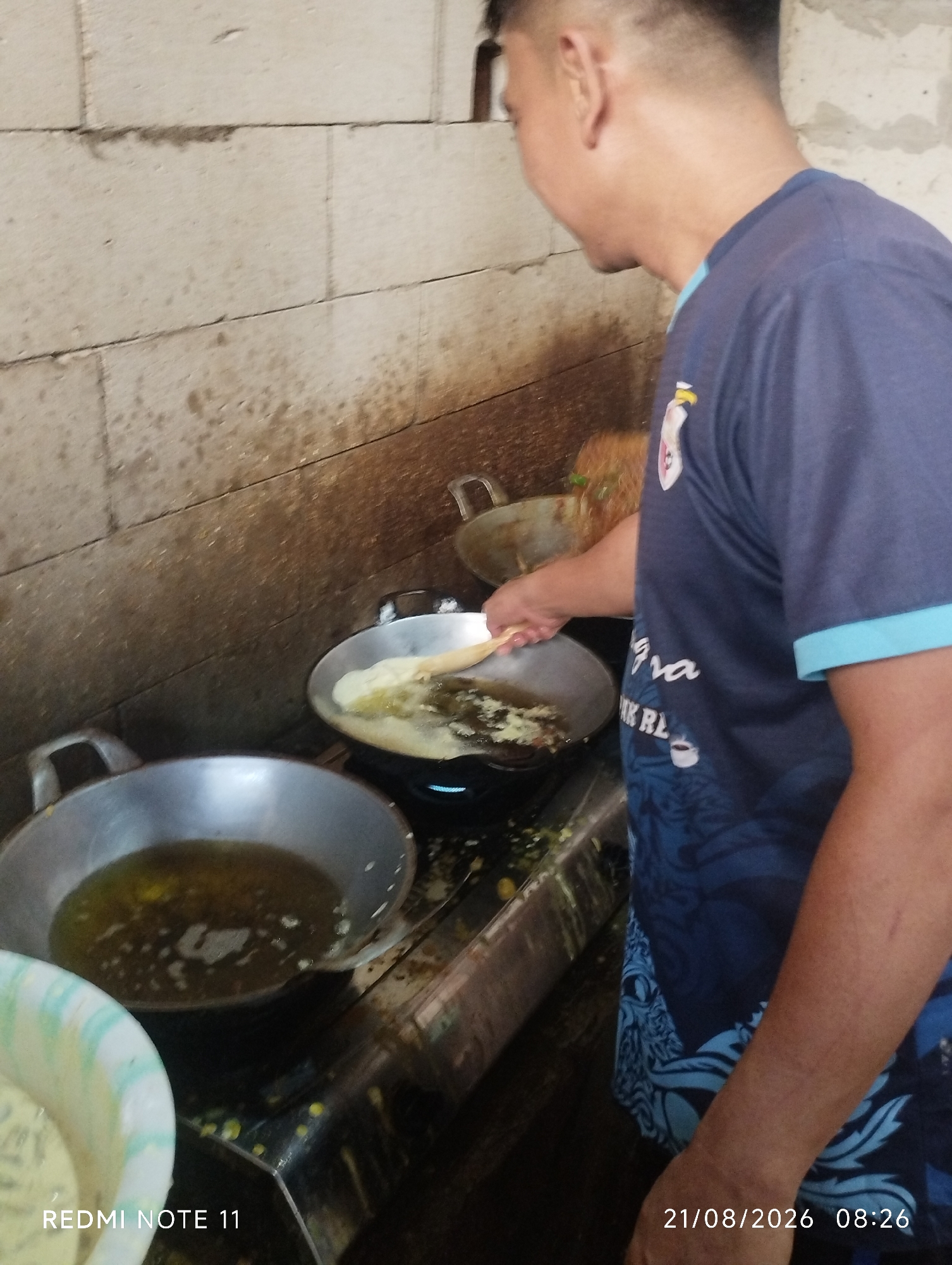 A man, viewed from the right side and from the shoulder up, is standing in front of a cooking area. He is wearing a dark blue t-shirt with a light blue trim on the collar and sleeves. His hair is short. He is looking down and to his left, with his right hand holding a wooden spatula, which is currently in a wok. The wok, which is on a stove, contains oil and what appears to be food being fried. To the left of this wok is another metal pan containing oil. In the foreground on the left, a portion of a white and green bowl is visible, containing a light-colored batter or liquid. The background is a wall constructed of light-colored cinder blocks, with some staining and discoloration. The cooking area appears to be in an outdoor or semi-outdoor setting, possibly a roadside stall. The image has a timestamp and text overlay in the bottom left and right corners, indicating "REDMI NOTE 11" and "21/08/2026 08:26".