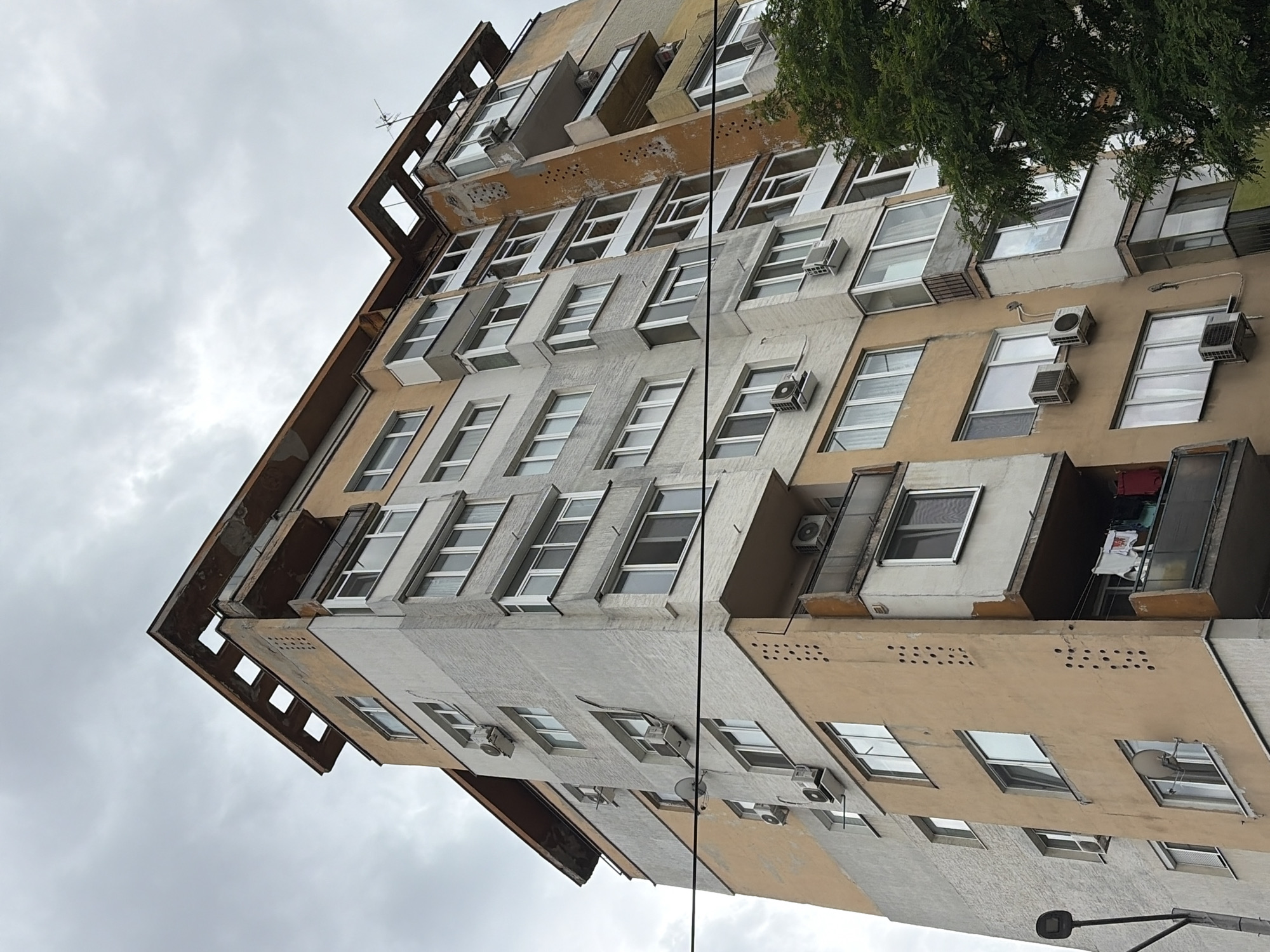 An exterior, low-angle, full shot captures the side of a weathered, multi-story apartment building against an overcast sky. The building exhibits a utilitarian architectural style, common in urban settings. Its facade is a patchwork of light gray and pale orange concrete, showing signs of age and wear, with peeling paint and some visible cracks. Numerous windows, mostly with white frames, punctuate the building's surface, some appearing as balconies with glass enclosures. Air conditioning units are visible mounted on many of the exterior walls. A satellite dish is attached to one of the lower floors. The top of the building features a distinctive, cantilevered roofline with open square sections. To the right of the building, a dense green tree provides a natural contrast to the urban structure. Power lines are strung horizontally across the lower portion of the image. A street lamp is visible on the far left. The scene is devoid of human presence.