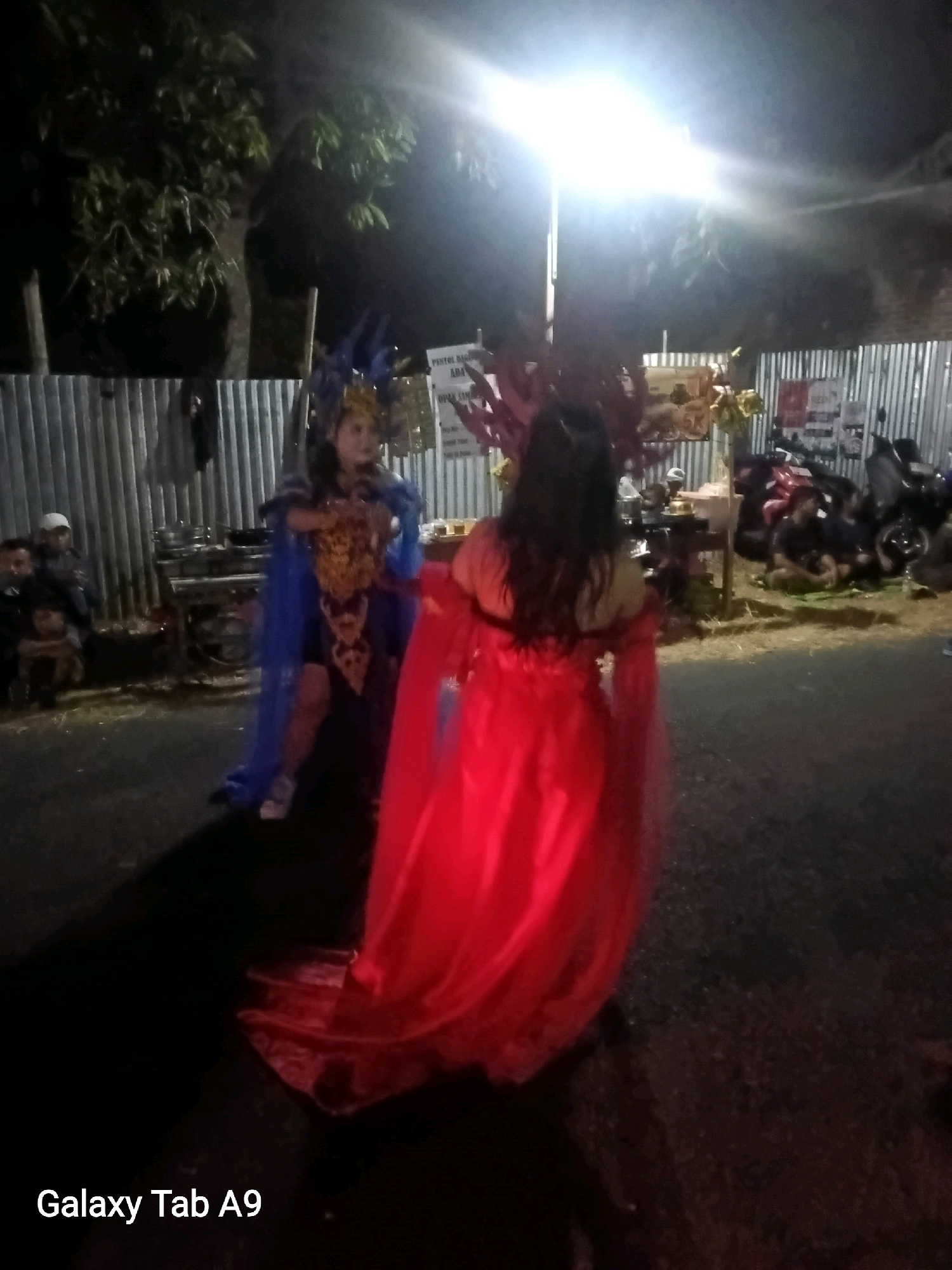 In the city of Wonoanti, Indonesia, two individuals are captured in a dimly lit outdoor scene. The person on the left, facing the camera, is adorned in a blue and gold traditional costume, complete with an elaborate headdress. To their right, another individual, viewed from the back, wears a flowing red gown. They are standing on a paved surface, with a corrugated metal fence forming a backdrop. Beyond the fence, motorbikes are parked, and several spectators, including a person in a white cap, are seated or standing, observing the event. A bright light source overhead illuminates the area, creating strong shadows. The atmosphere suggests a cultural performance or celebration taking place in Wonoanti.
