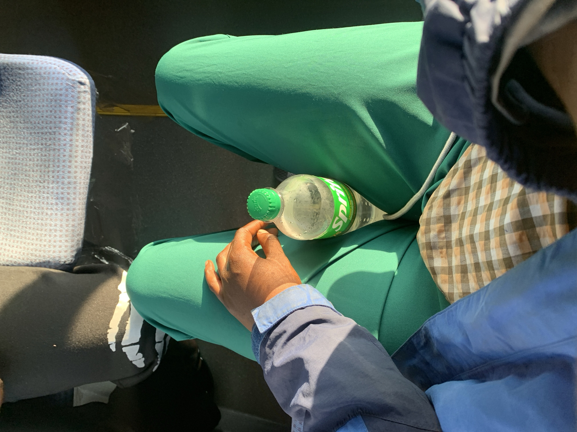 The image shows a person sitting in a vehicle, likely a bus, in Rafin Par, Nigeria. The person is wearing green scrubs and a blue jacket. A bottle of Sprite is placed on their lap. The background is blurry, but it appears to be the interior of a bus with patterned seats. The lighting suggests it is daytime.