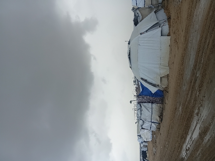 The image displays a temporary encampment in Khan Yunis, Palestinian Territory, under a heavily overcast sky dominated by dark grey clouds. The ground is unpaved, consisting of muddy sand with visible tire tracks and puddles, indicating recent rain. A large, multi-faceted dome-shaped tent, primarily light-colored and appearing soiled at its base, occupies the foreground on the right, featuring visible structural supports. To its left and extending into the background is a series of smaller, predominantly rectangular tents, also constructed from light-colored fabric. Some of these smaller shelters incorporate sections of blue tarpaulin. The encampment continues into the distance, with faint structures resembling fences or barriers visible in the far background. No individuals or specific actions are discernible within the frame.