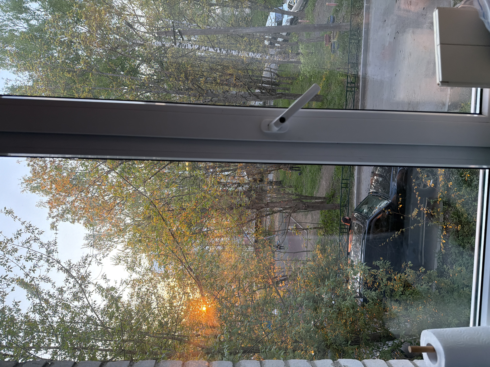 View from a window overlooking a street in Tokkari-Lend, Russia. A black car is parked near the window. Trees line the street, with the setting sun visible through the leaves. There is some precipitation on the street in the distance. A roll of paper is near the window.
