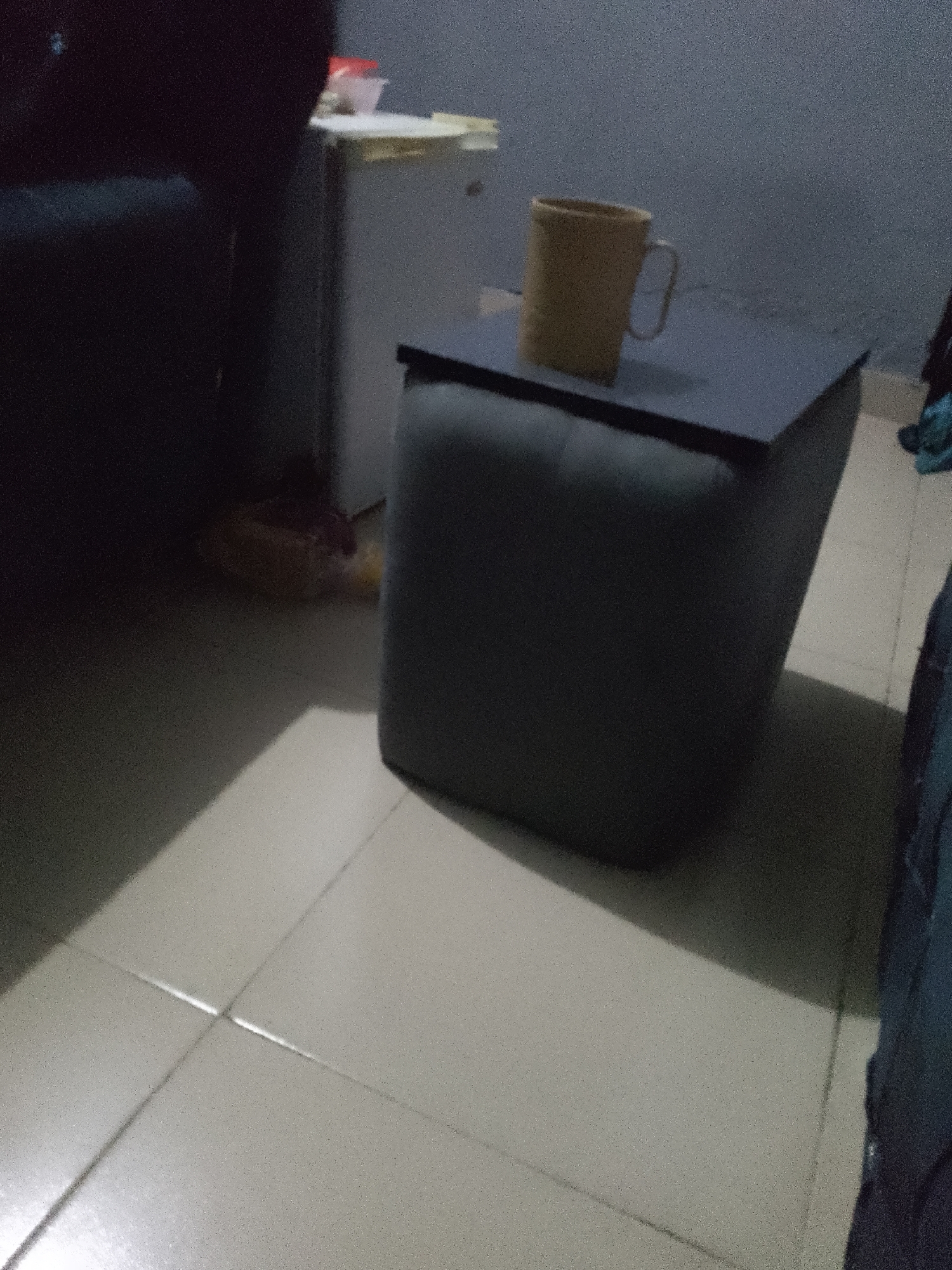 A dimly lit indoor scene, possibly a domestic or office environment, is depicted. The foreground shows light-colored tiled flooring with distinct grout lines. A large, dark, cubical ottoman or storage unit is centrally positioned. On top of this unit rests a dark rectangular surface, resembling a tabletop. A single, light-brown mug with a handle is placed on this tabletop, slightly off-center. To the left of the ottoman, a dark, out-of-focus object, possibly furniture, dominates the background. Behind the ottoman, a light-colored appliance, likely a mini-fridge, is partially visible. A plastic bag is seen on the floor near the appliance. The overall lighting suggests a low-light condition, with shadows cast on the floor, indicating a light source from the right. The scene appears static, with no people or overt actions taking place. There are no discernible elements to indicate a specific location in the city of Jalingo, Nigeria.