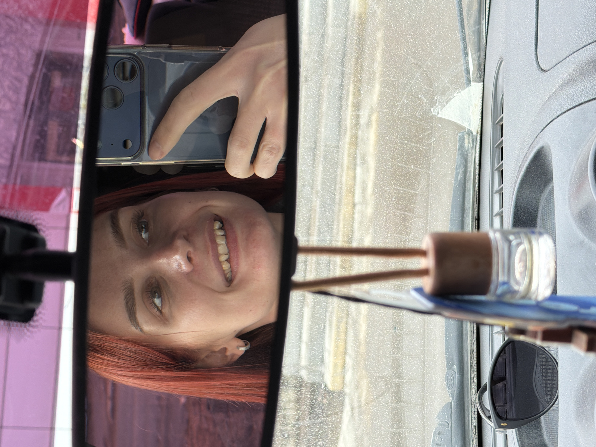 The image depicts a woman taking a selfie in the rearview mirror of a vehicle. The woman has red hair and is smiling. Her hand is holding a smartphone up to the mirror, obscuring part of her face. The rearview mirror is positioned in front of a dirty windshield. Visible outside the windshield is an urban environment with a gray/white exterior. A small air freshener, consisting of a glass container with a brown top and two wooden sticks, is located near the mirror. The location is identified as Chaikovsky, Russia.