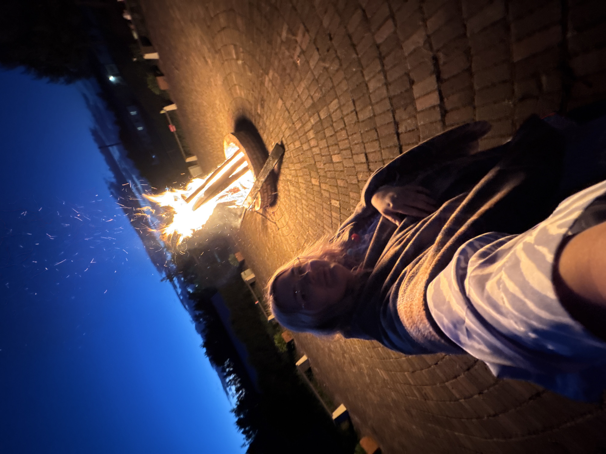 The image shows a person standing in front of a bonfire at dusk. The person is wrapped in a blanket and appears to be taking a selfie. The bonfire is in a circular fire pit, and sparks are flying up into the night sky. The ground is paved with bricks, and there are trees and buildings in the background. The overall scene suggests a relaxed and cozy atmosphere. The location is Birobidzhan, Russia, as stated.