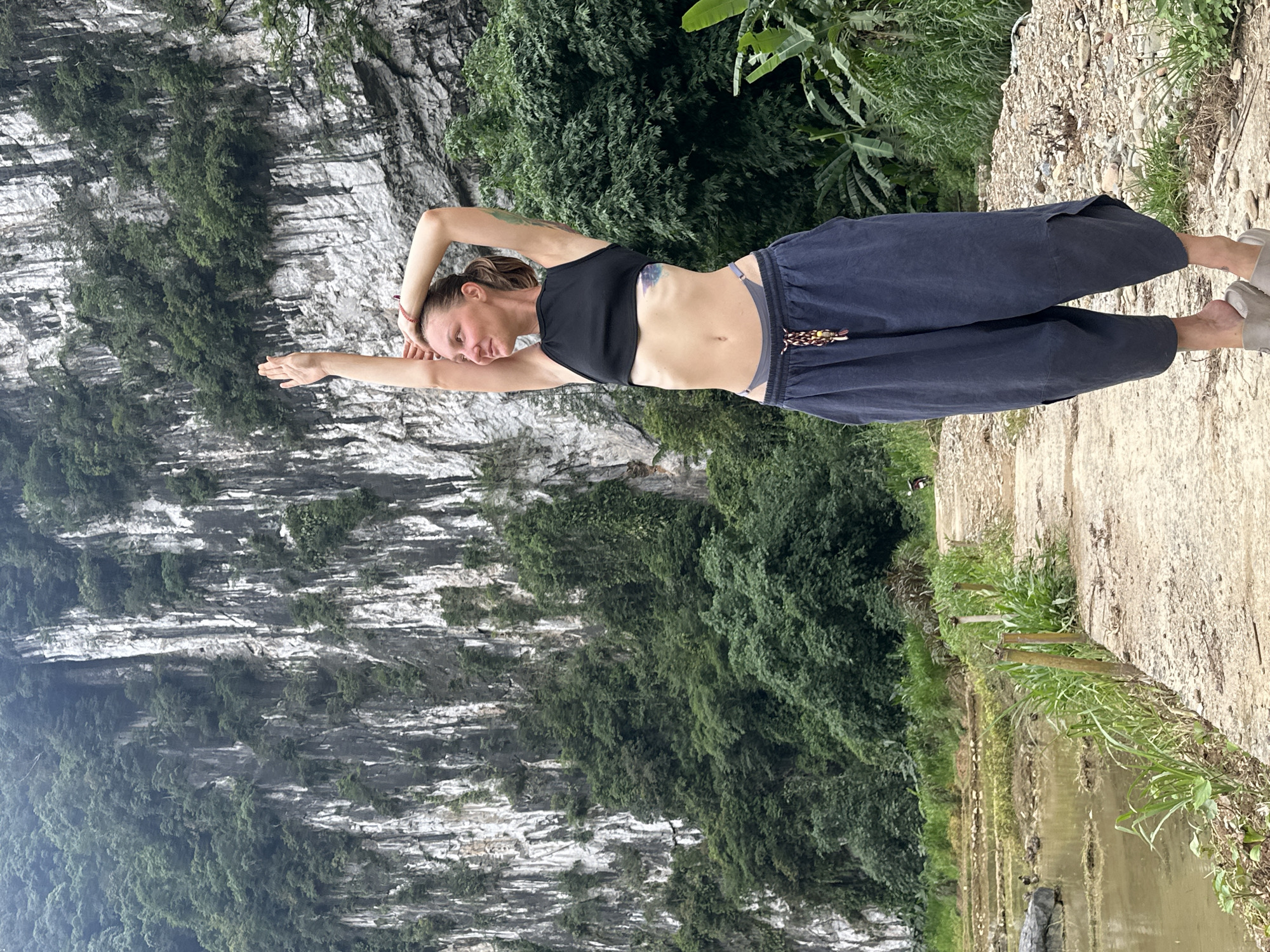 A woman in a black crop top and dark blue pants stands on a dirt path with her arms raised above her head. She has a tattoo on her left side. Behind her is a steep, rocky cliff face covered in dense green foliage. To her left are rice paddies with calm, muddy water and some small dark birds. The path continues into the distance, bordered by vegetation. The overall setting appears to be a natural, rural landscape in Pù Luông Commune, Vietnam.