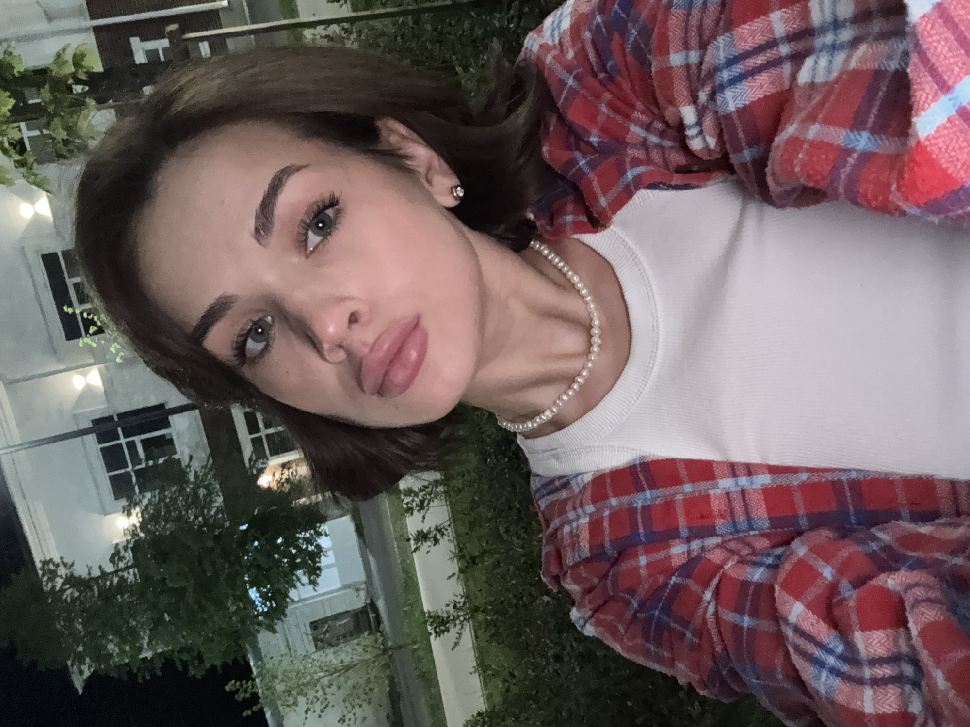 A young woman with brown hair, wearing a white top and a red and blue plaid flannel shirt, is taking a selfie outdoors at night. She is also wearing a pearl necklace and small earrings. The background features a white building with multiple windows and greenery, suggesting a residential or commercial area in Sarapul, Russia, based on the image's origin. The lighting is artificial, indicating it is nighttime.